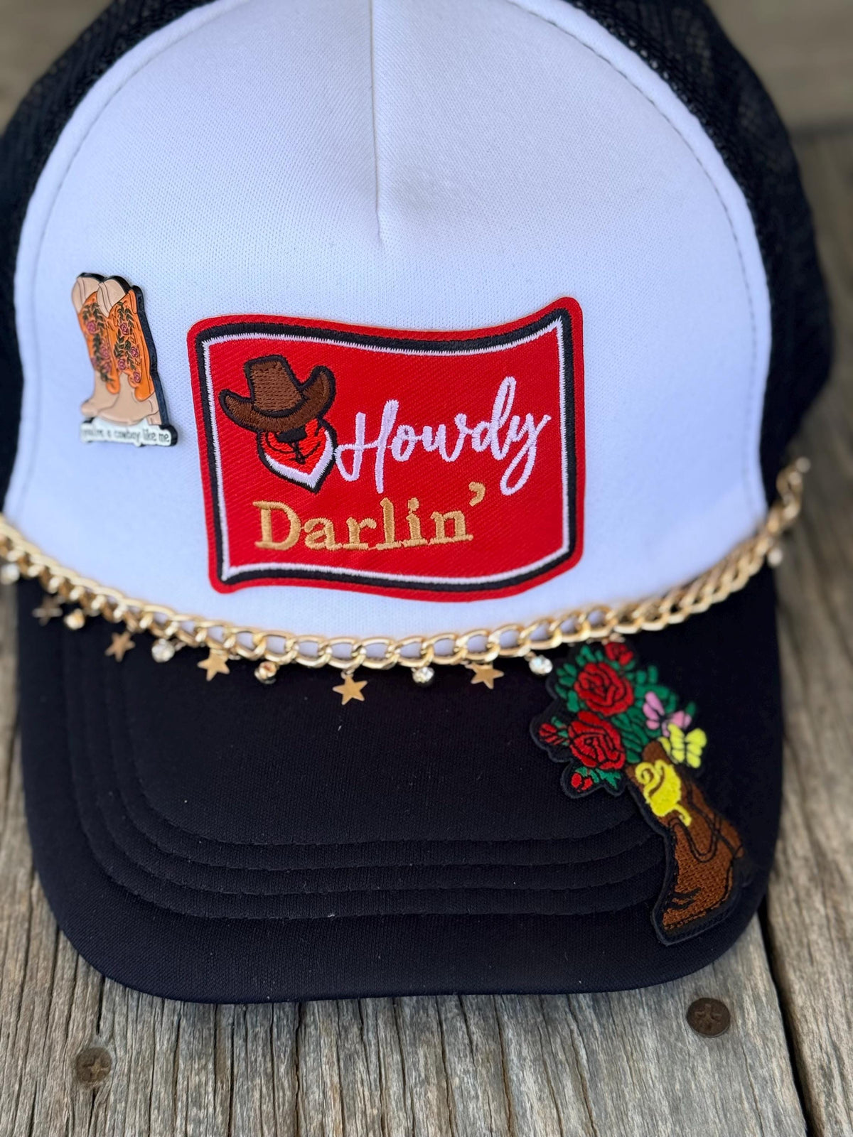 A white and black trucker hat with a red embroidered patch reading 'Howdy Darlin'', decorated with a gold chain, cowboy boot patch, and floral boot patch, shown on a rustic wooden surface.