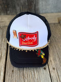 A black and white trucker hat with a red 'Howdy Darlin'' patch on the front, decorated with a gold chain and cowboy boot patches, resting on a rustic wooden surface.