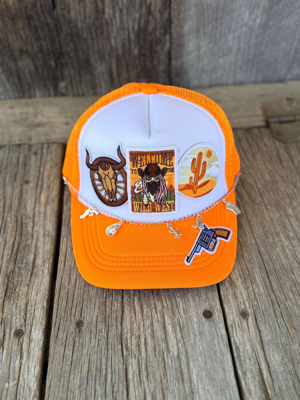 An orange and white trucker hat with distressed details, decorated with three western-themed embroidered patches, displayed on a rustic wooden surface.