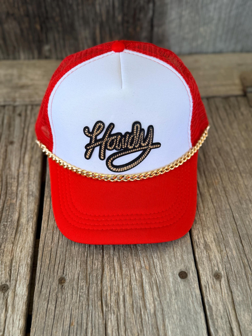 A red and white trucker hat with a gold chain detail across the front and 'Hardy' embroidered in black and gold on the white front panel, displayed on a rustic wooden surface.