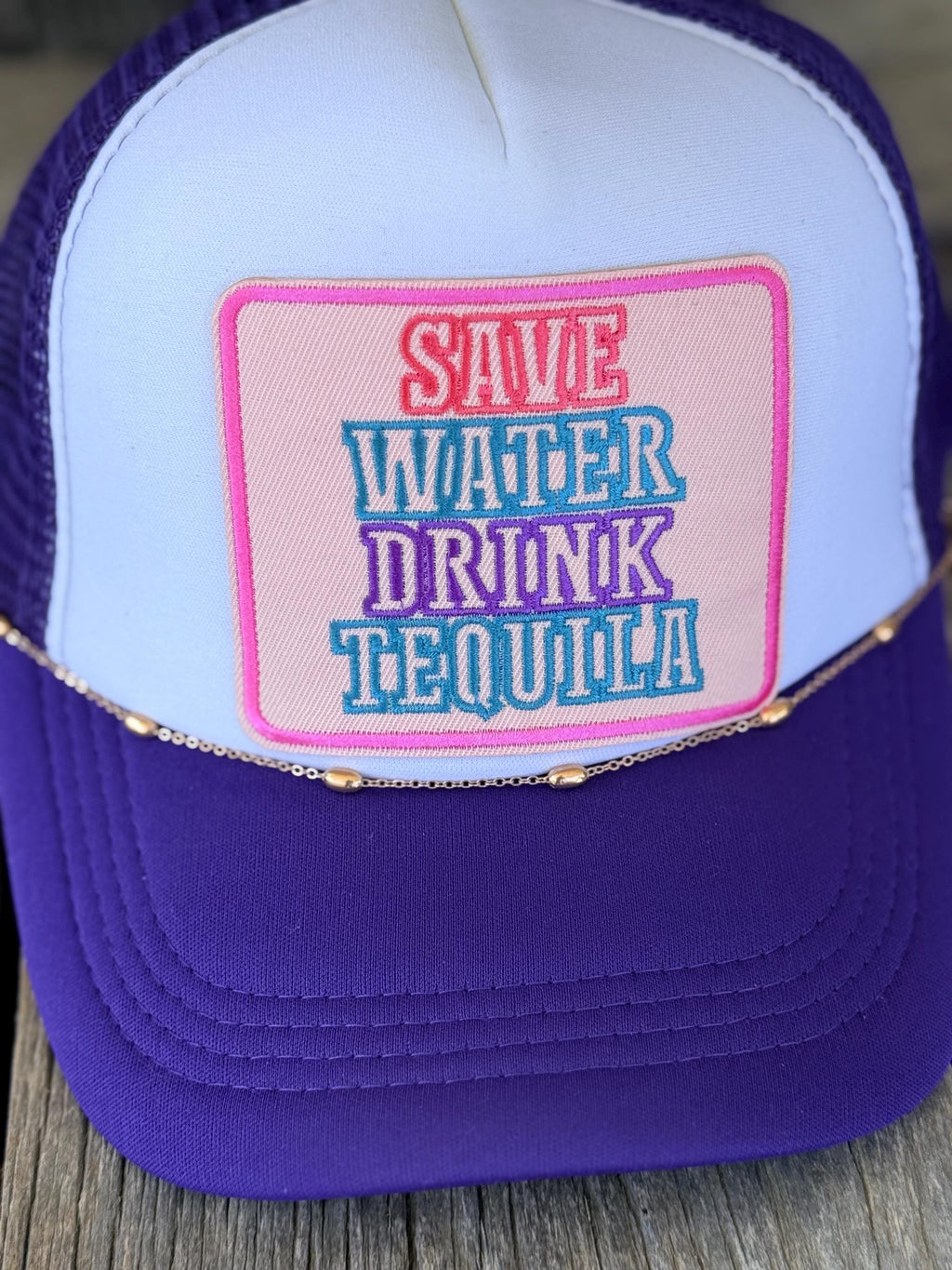A purple and white trucker hat with a pink patch that reads 'SAVE WATER DRINK TEQUILA' in colorful embroidered text, resting on a wooden surface.