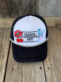 A black and white trucker hat with a silver chain detail across the front, featuring a custom patch with red cherries and a blue cowboy illustration, displayed on a rustic wooden surface.