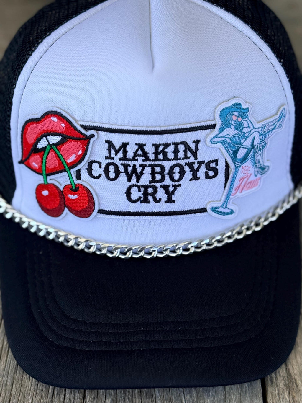 A black and white trucker hat with a silver chain across the front. The front panel is white with embroidered patches including red cherries with lips and a blue cowboy graphic.