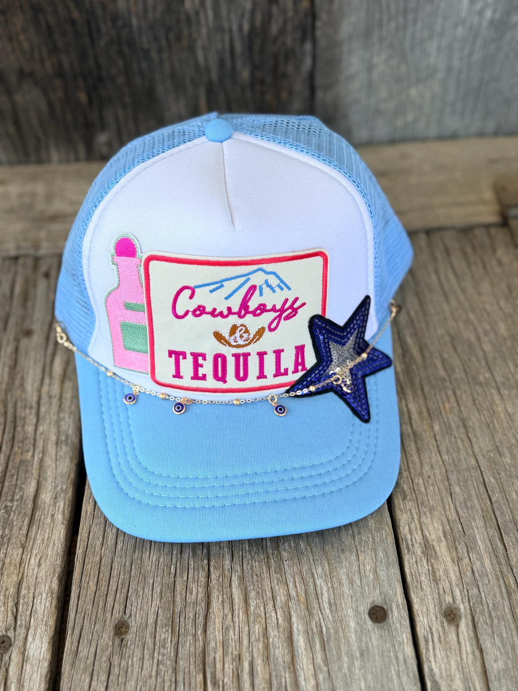A light blue and white trucker hat with a 'Cowboys & Tequila' patch, resting on a rustic wooden surface.