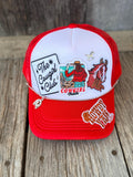 A red and white trucker hat with western-themed embroidered patches, including 'The Cowgirl Club', a cowboy, a horse, and 'Cowgirl Trails', displayed on a rustic wooden surface.