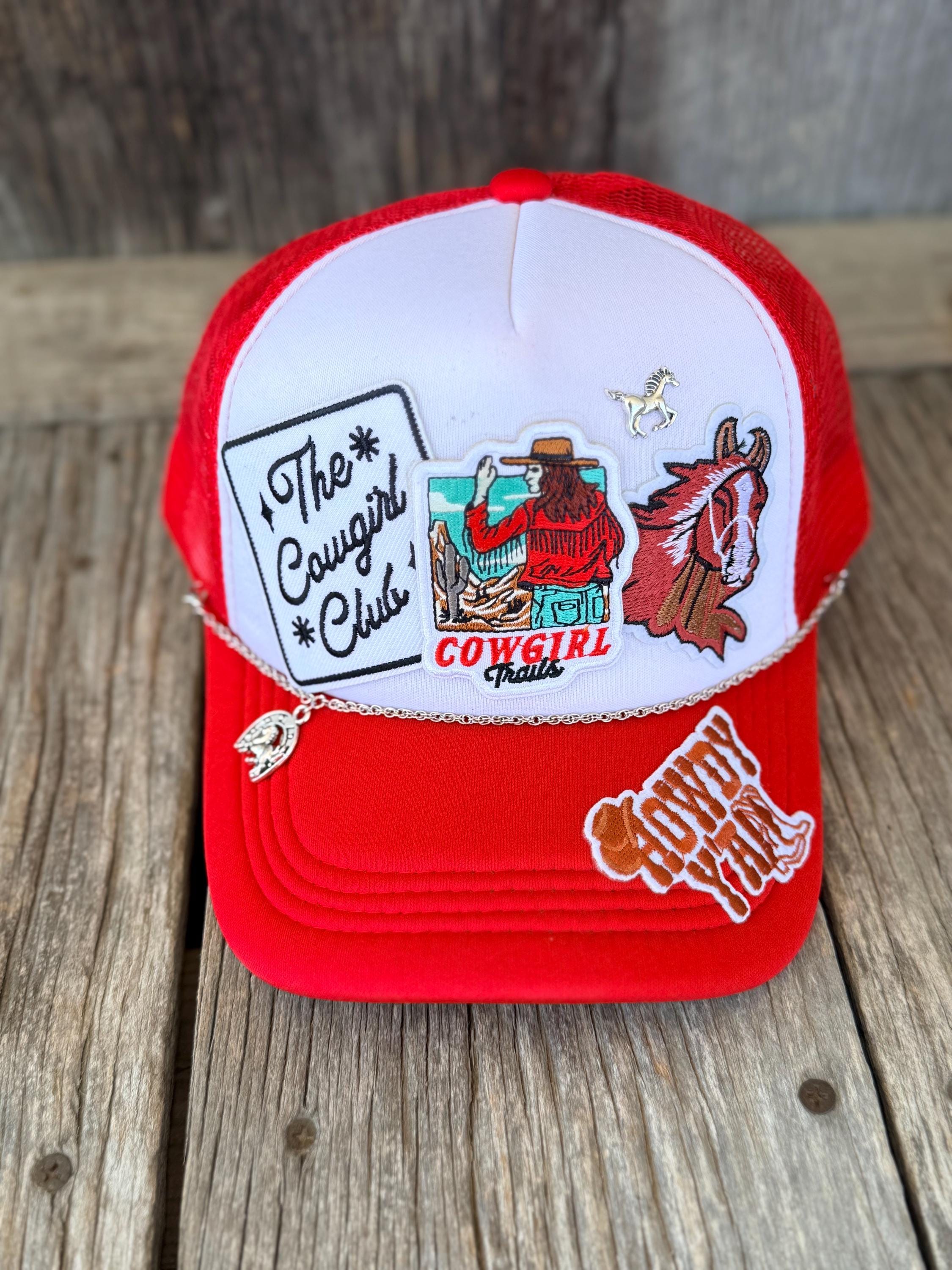 A red and white trucker hat with western-themed embroidered patches, including 'The Cowgirl Club', a cowboy, a horse, and 'Cowgirl Trails', displayed on a rustic wooden surface.