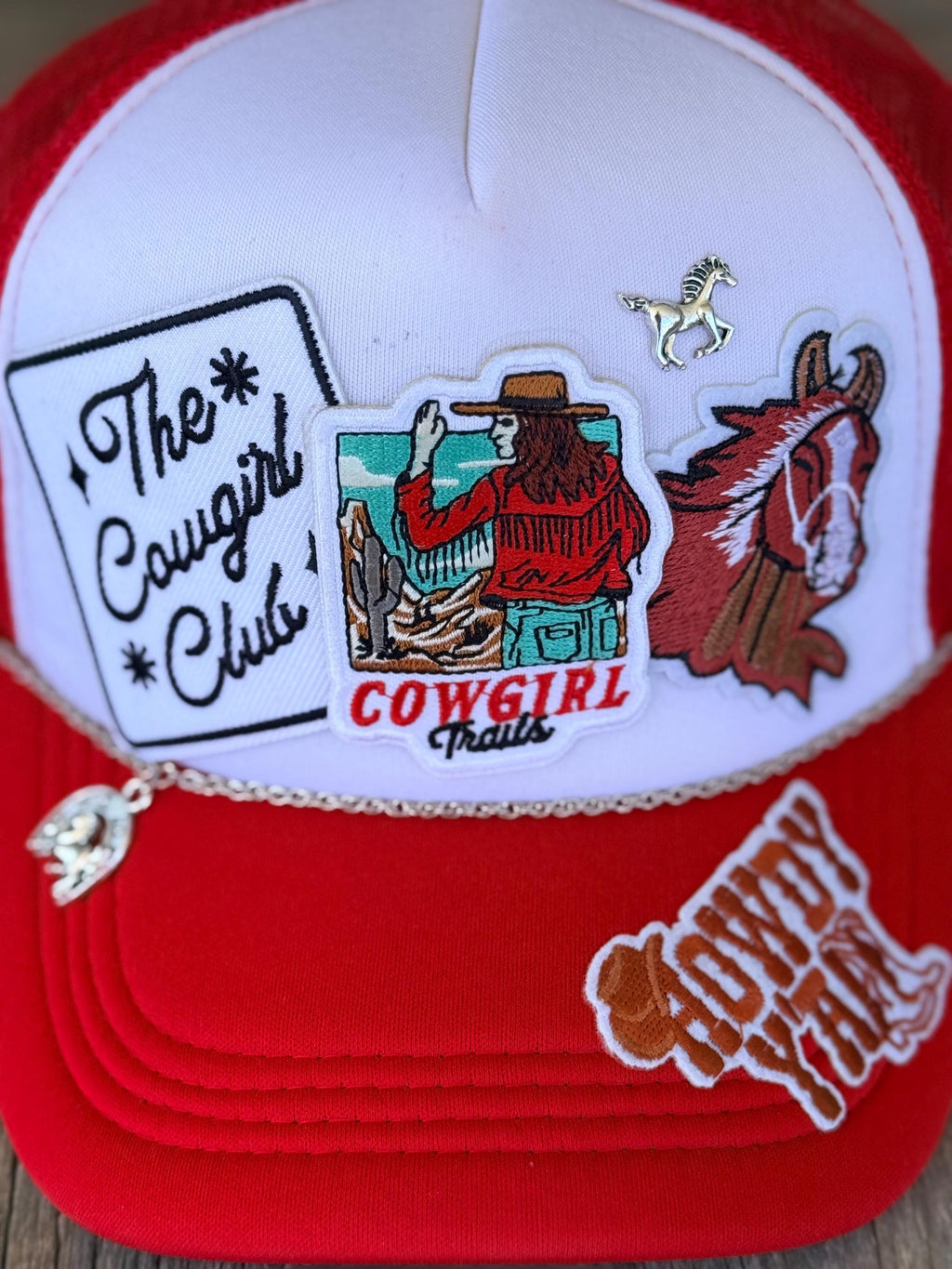 A red and white trucker hat decorated with multiple embroidered patches, including 'The Cowgirl Club', 'Cowgirl Trails', and a cowboy riding a horse, with a small silver horse charm hanging from the brim.