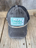 A distressed dark gray trucker hat with a black mesh back, featuring a front patch that reads 'Beautiful CRAZY' in teal script and block lettering on a light blue background. The hat is displayed on a rustic wooden plank surface.