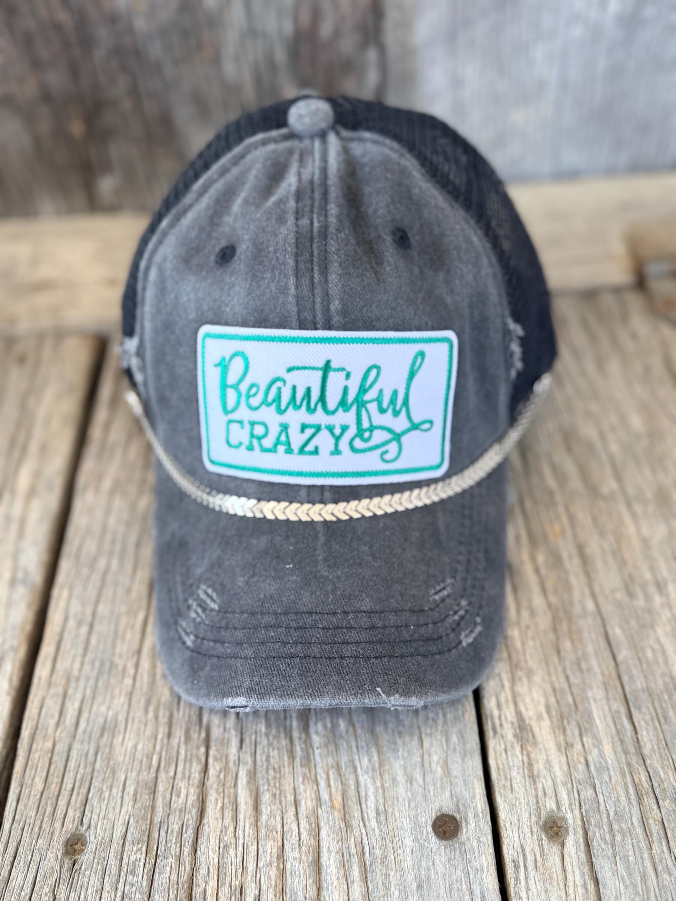 A distressed dark gray trucker hat with a black mesh back, featuring a front patch that reads 'Beautiful CRAZY' in teal script and block lettering on a light blue background. The hat is displayed on a rustic wooden plank surface.