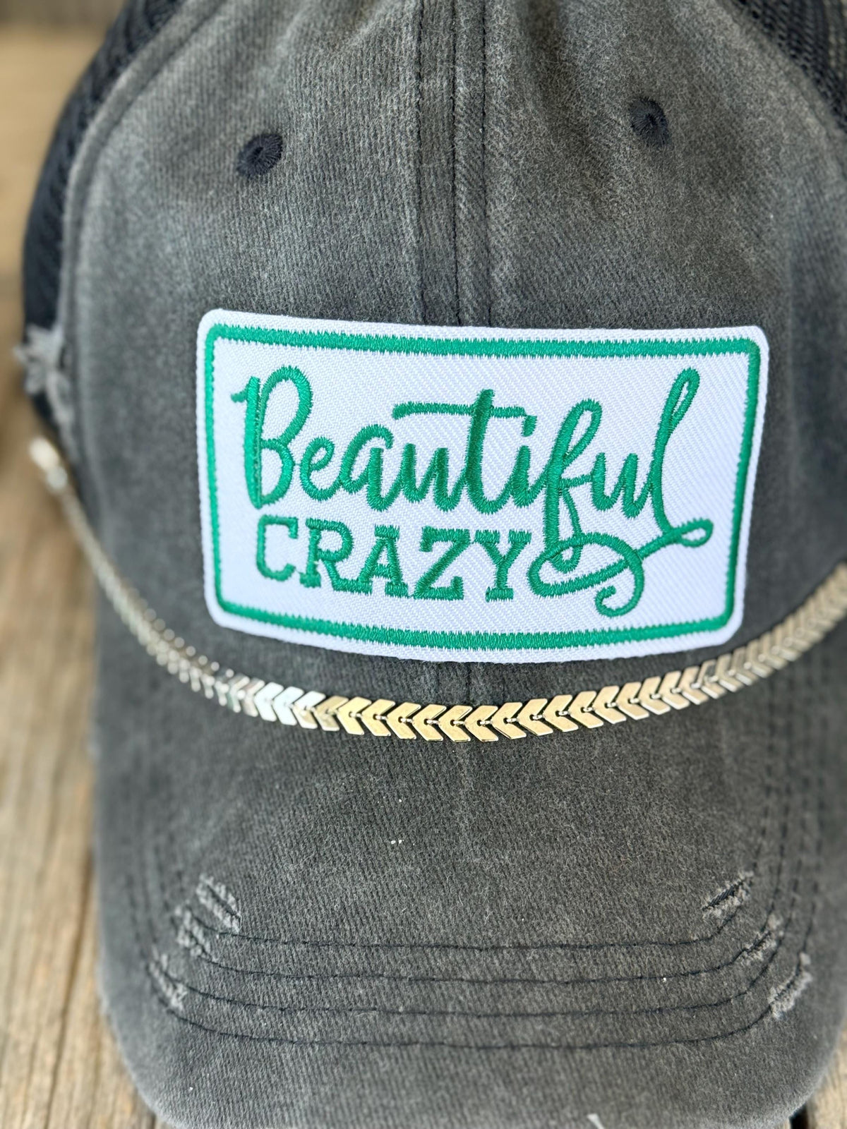 A close-up of a distressed dark gray trucker cap featuring a white rectangular patch with green embroidered text that reads 'Beautiful CRAZY'.