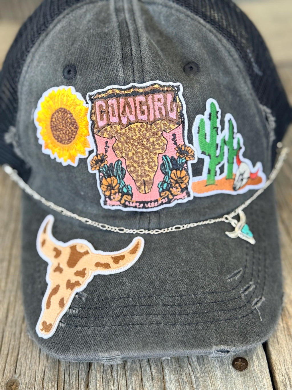 A distressed dark gray trucker hat with a western-themed embroidered patch on the front, a longhorn patch on the brim, and a silver chain with a small heart charm resting across the brim.