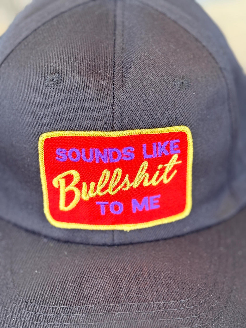 A close-up of a dark gray baseball cap featuring a red rectangular embroidered patch with yellow and blue text that reads 'SOUNDS LIKE Bullshit TO ME'.