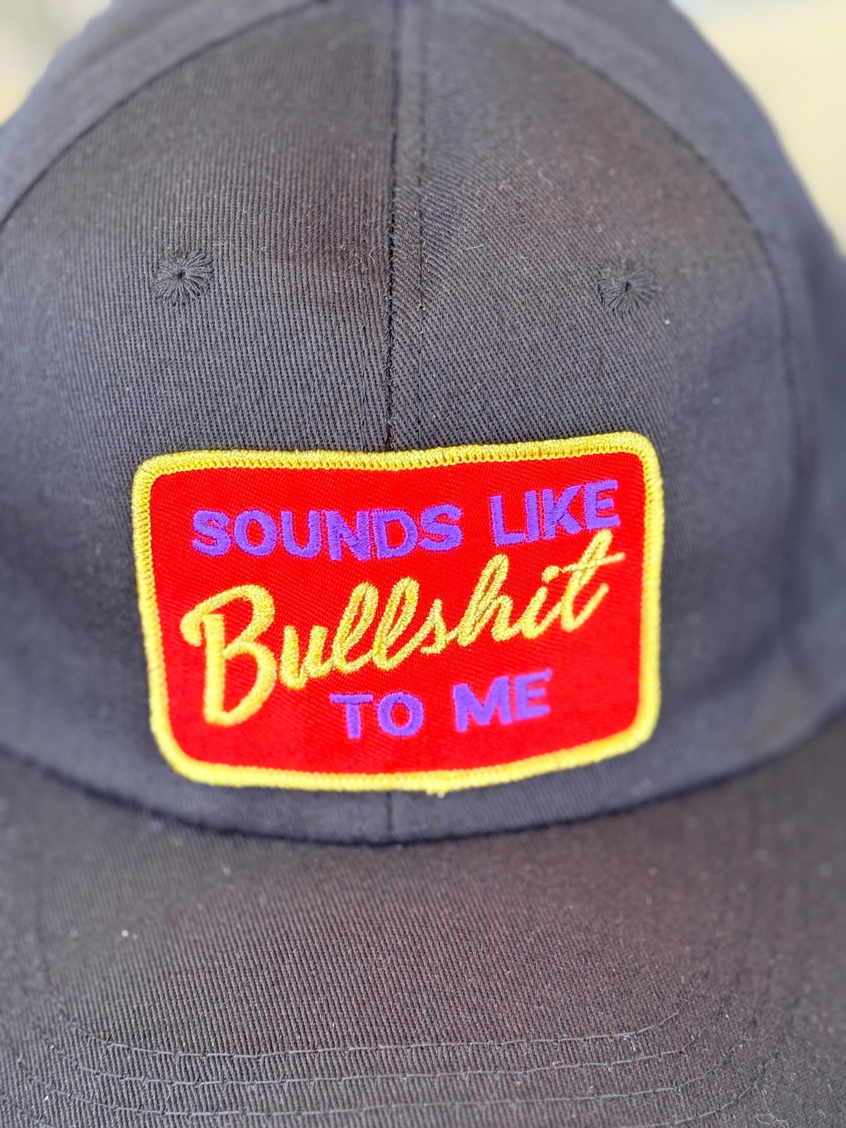 A close-up of a dark gray baseball cap featuring a red rectangular embroidered patch with yellow and blue text that reads 'SOUNDS LIKE Bullshit TO ME'.