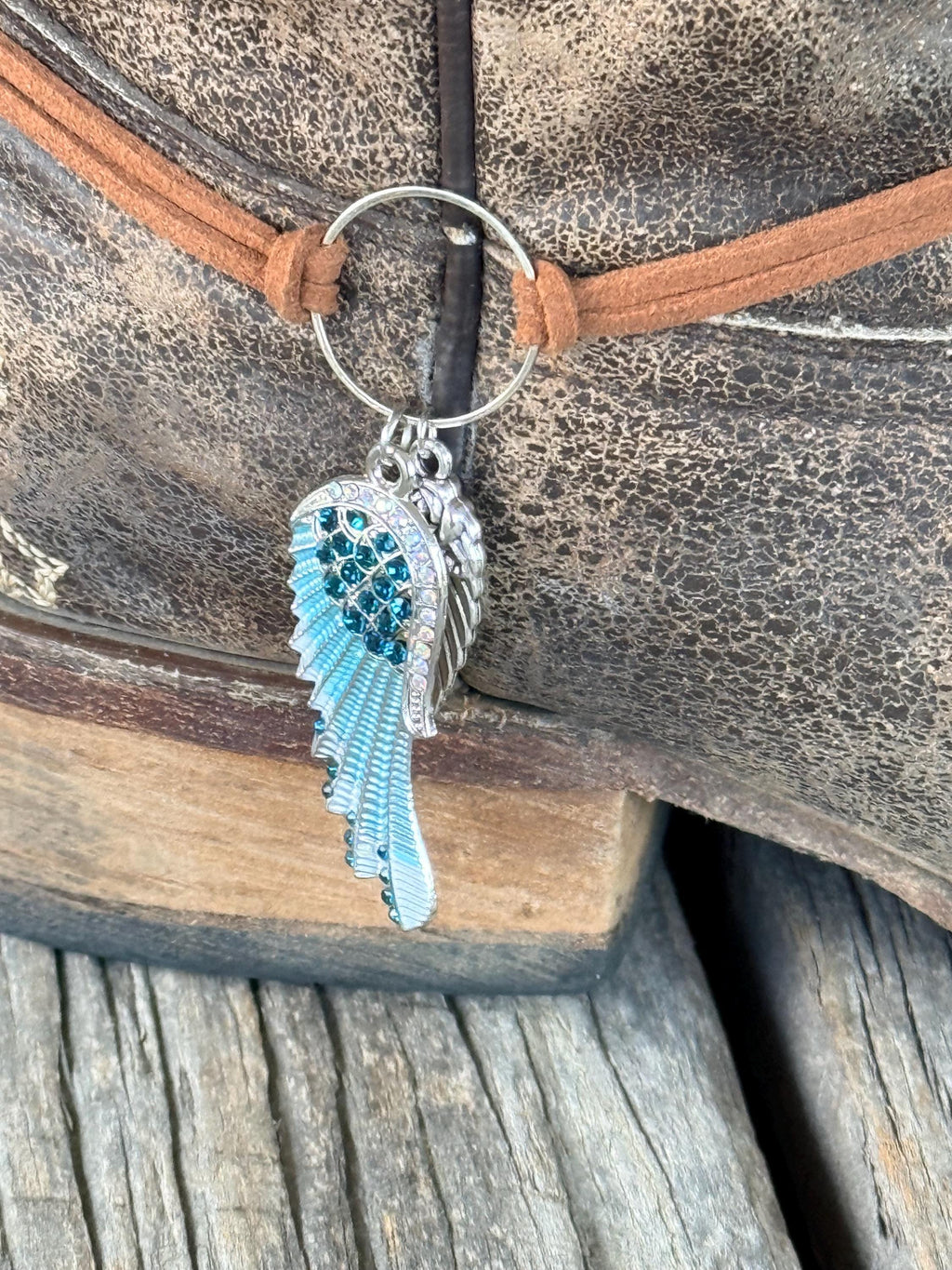 Macro close-up of two-piece angel wing boot bracelet charm with silver engraved wing and turquoise crystal-encrusted wing with teal rhinestones, silver ring connector, and brown leather cord, Western boho boot jewelry