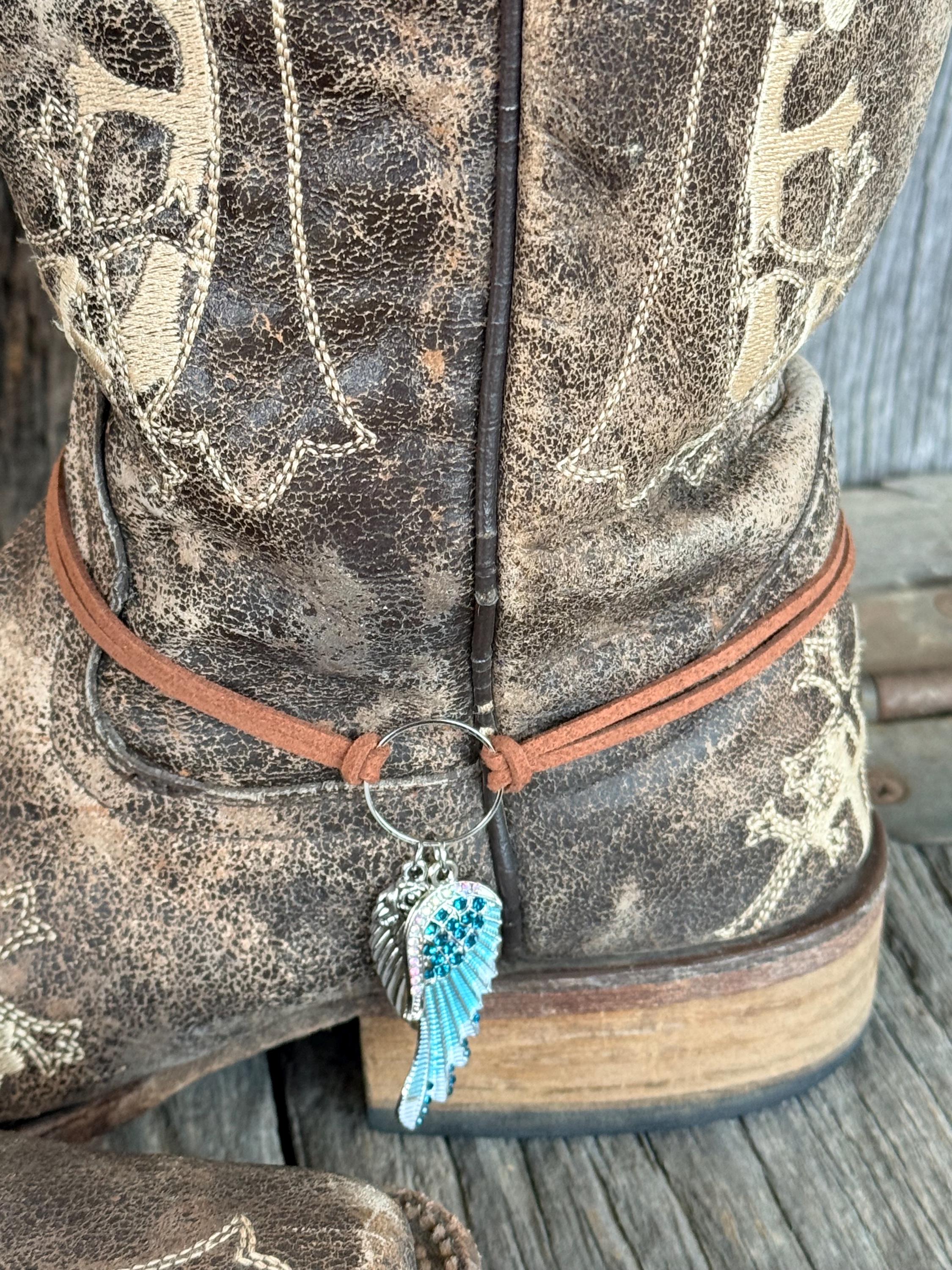 Back heel close-up of brown distressed cross-embroidered cowboy boot with thin brown leather cord boot bracelet and two-piece angel wing charm featuring silver engraved wing and turquoise crystal-encrusted wing with teal rhinestones