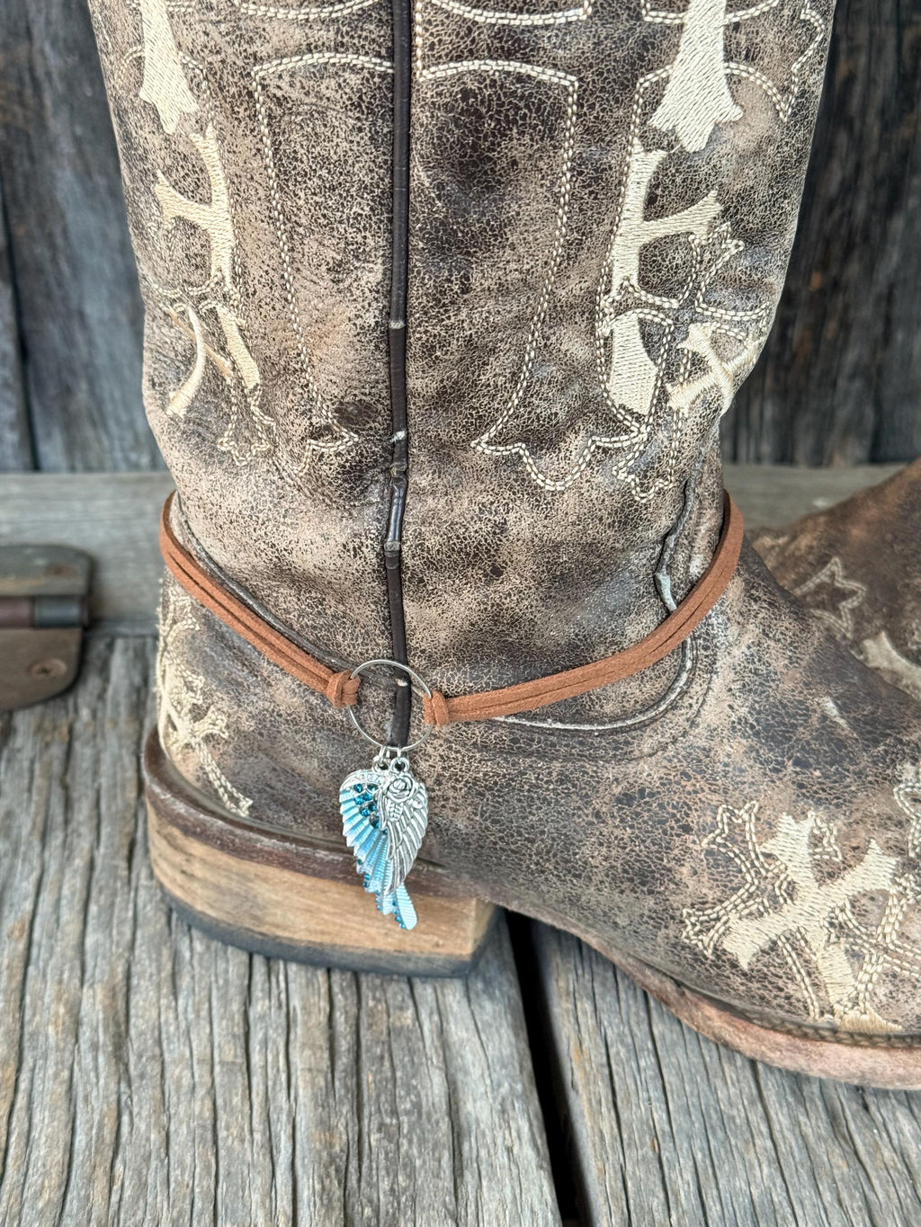 Front ankle close-up of brown distressed cross-embroidered cowboy boot with thin brown leather cord boot bracelet and two-piece silver and turquoise crystal angel wing charm pendant