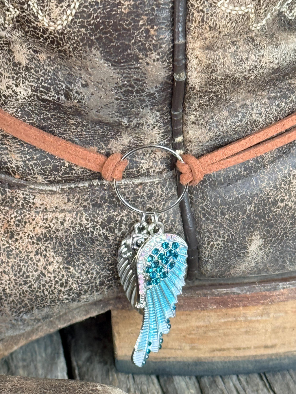 Macro close-up of two-piece angel wing boot bracelet charm showing silver engraved wing and turquoise crystal-encrusted wing with teal rhinestones detail, on brown leather cord with silver ring connector