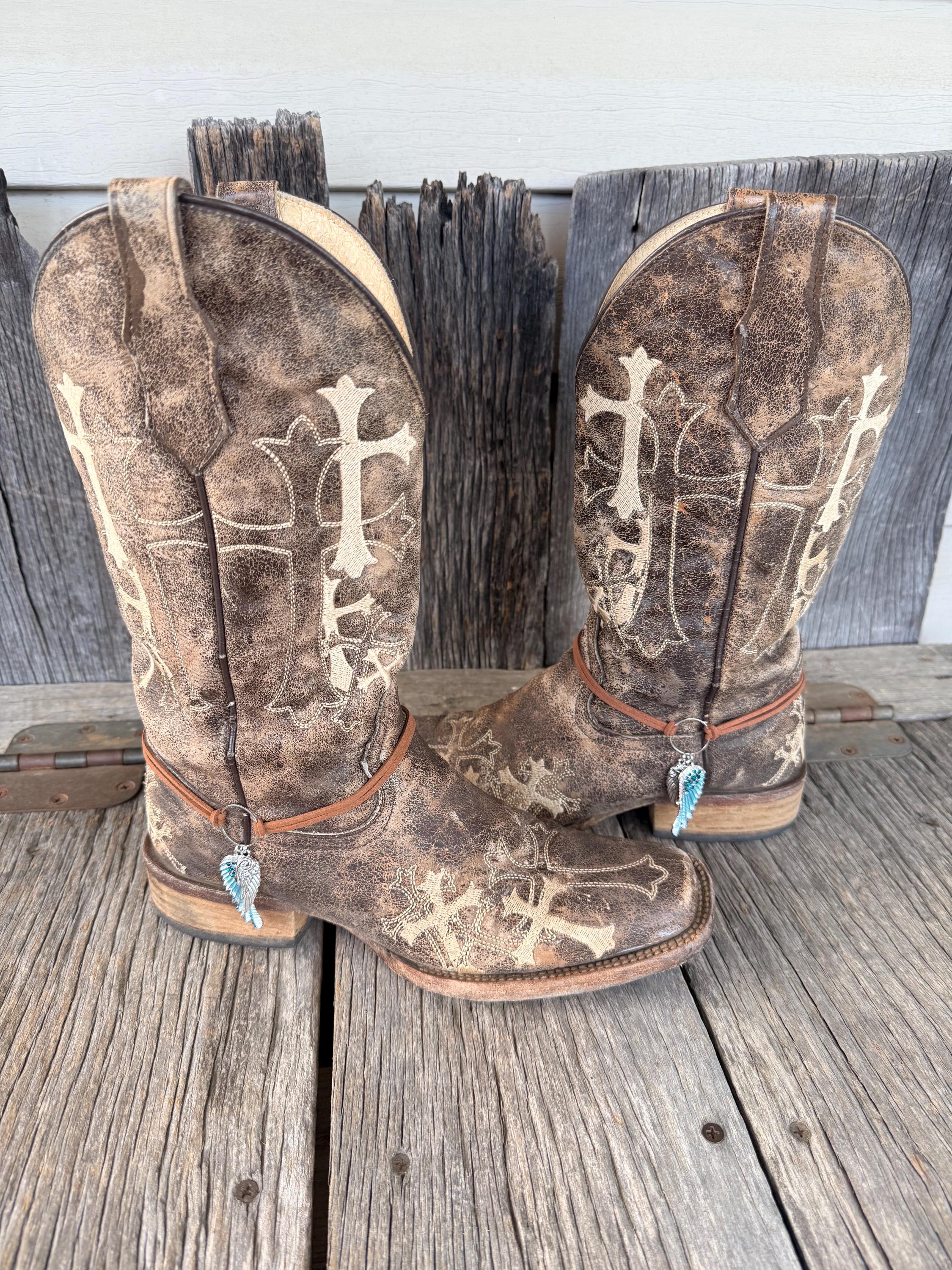 Front view of pair of brown distressed cross-embroidered cowboy boots on weathered wood planks wearing thin brown leather cord boot bracelets with two-piece silver and turquoise crystal angel wing charm, Western boho boot jewelry set of 2