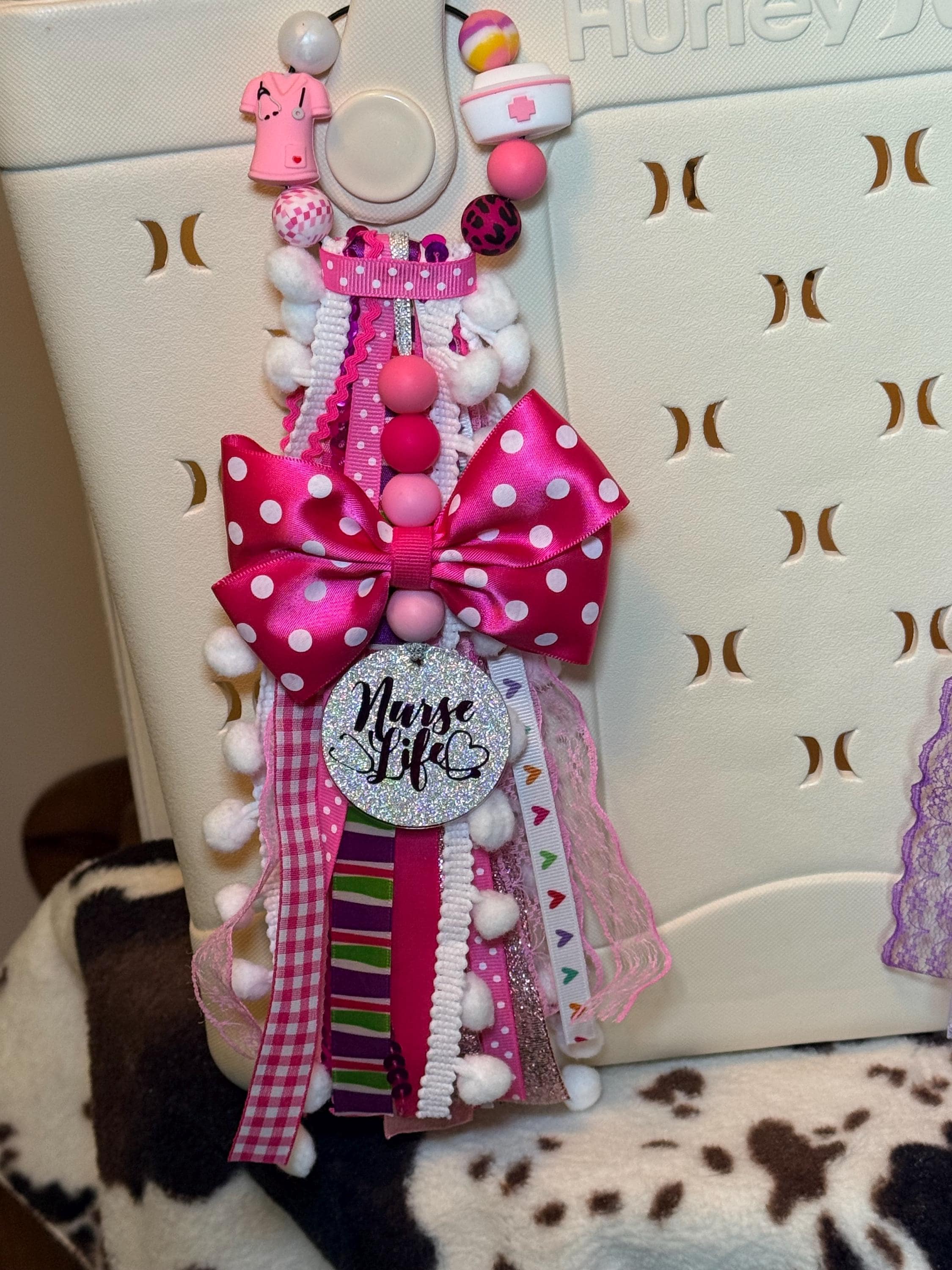 Pink Nurse Life silicone beaded bag tassel clipped to white Hurley tote bag handle on cowhide rug, featuring white pom-pom trim bead strand with mixed pink nurse-themed silicone beads including scrubs charm and nurse hat charm, hot pink polka-dot grosgrain bow, cascading multi-ribbon tassel in pink polka-dot gingham striped ric-rac lace glitter and heart-print ribbons, and silver glitter Nurse Life round pendant