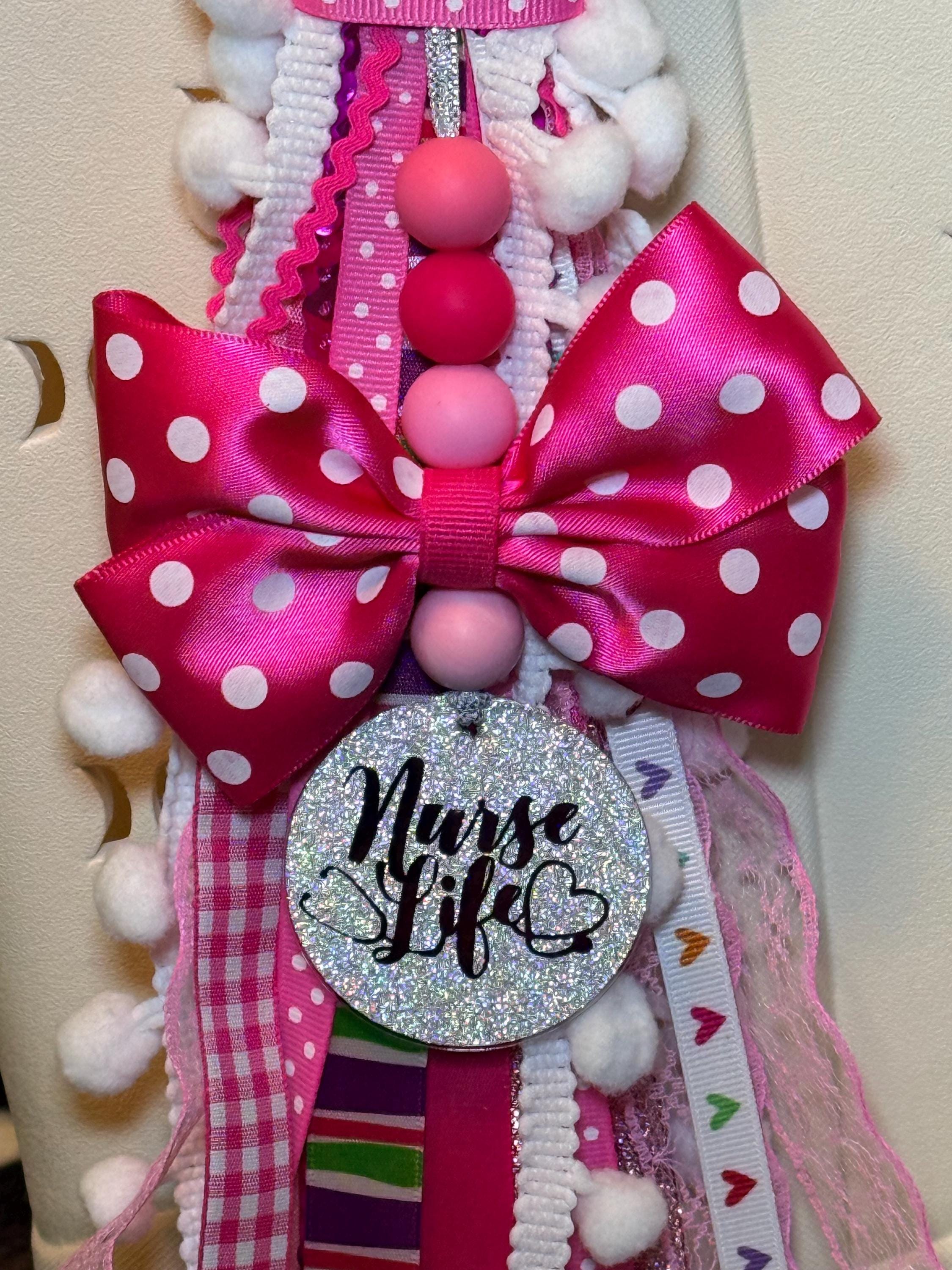 Close-up of pink Nurse Life bag tassel showing large hot pink polka-dot grosgrain bow with pink silicone beads, silver glitter round Nurse Life pendant with stethoscope heart detail hanging below bow, and cascading multi-ribbon tassel in pink polka-dot gingham striped ric-rac lace glitter and heart-print ribbons