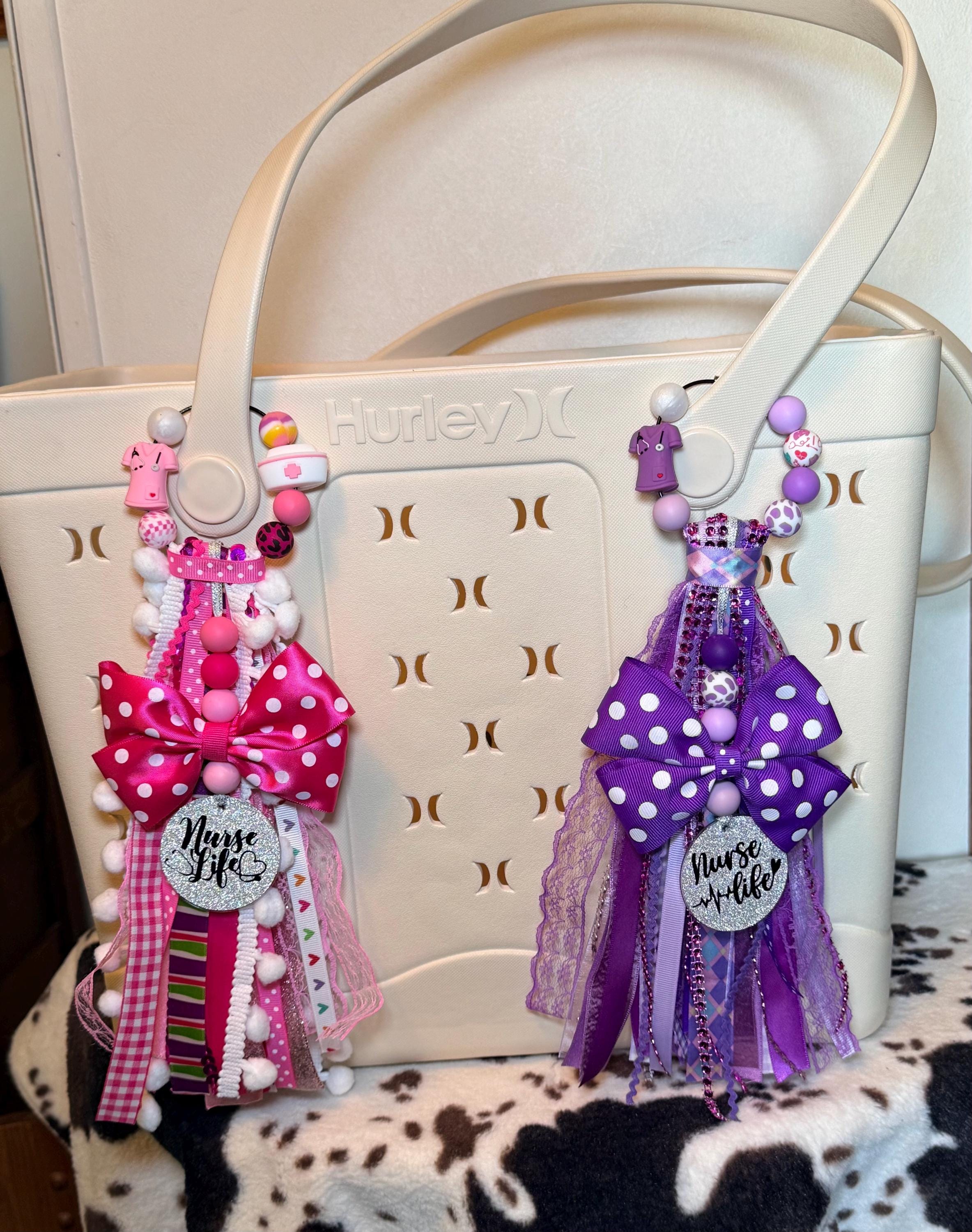 A cream-colored Hurley tote bag with decorative nurse-themed bag tassel charms hanging from the handles. The left charm is pink and white with a pink polka dot bow, and the right charm is purple and white with a purple polka dot bow.