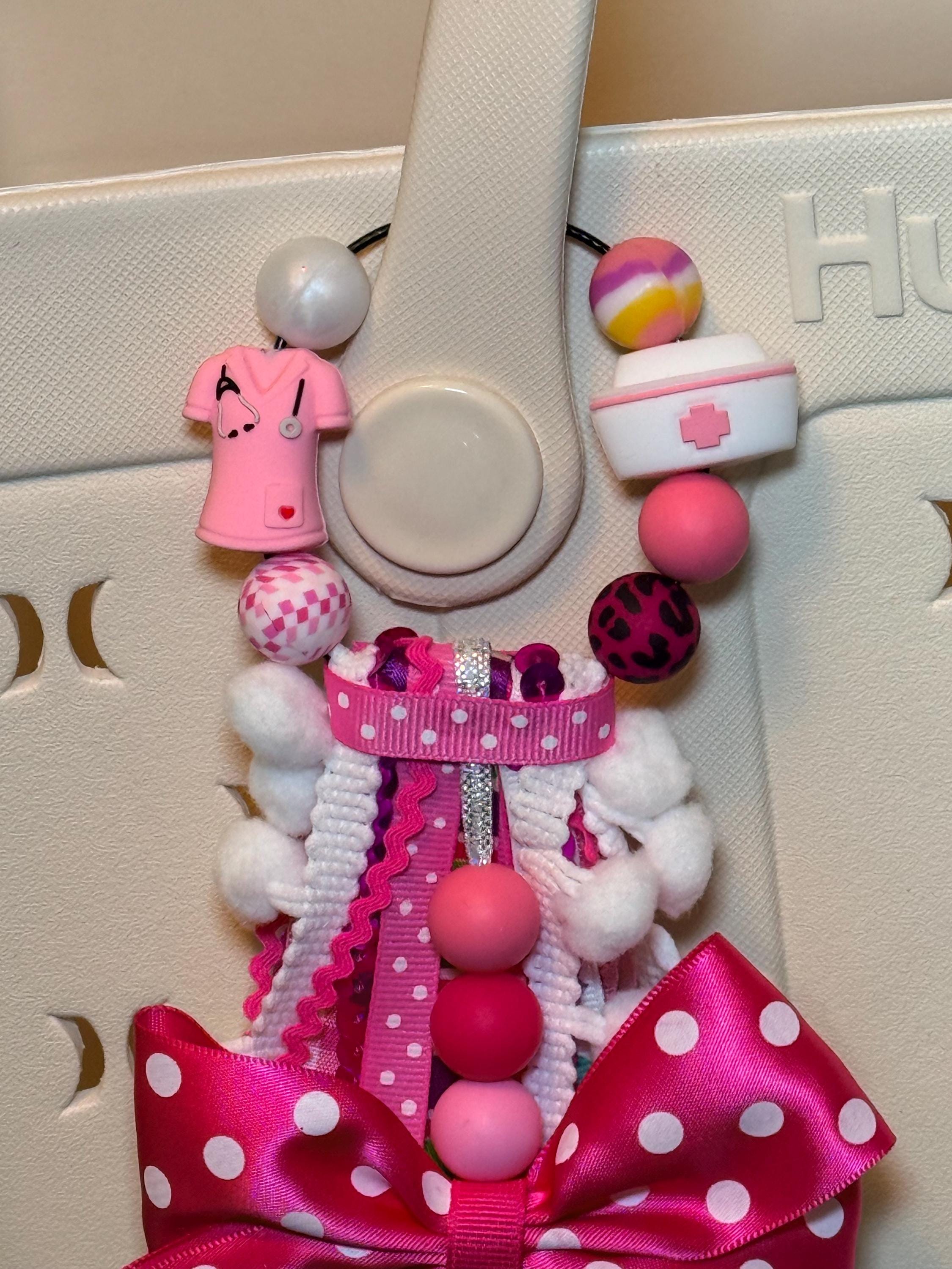 Close-up of pink Nurse Life bag tassel top showing wire loop clip with pink scrubs charm with stethoscope, nurse hat charm, candy-swirl bead, leopard print bead, checkered bead, and solid pink silicone beads over white Hurley tote bag handle, with hot pink polka-dot grosgrain bow and ribbon tassel top visible below