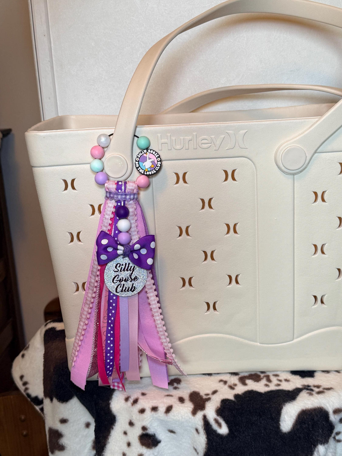 Silly Goose On The Loose Club silicone beaded bag tassel with glitter Silly Goose Club charm disc, purple polka dot bow, and layered pink and purple ribbon tassel clipped onto a white tote bag handle

