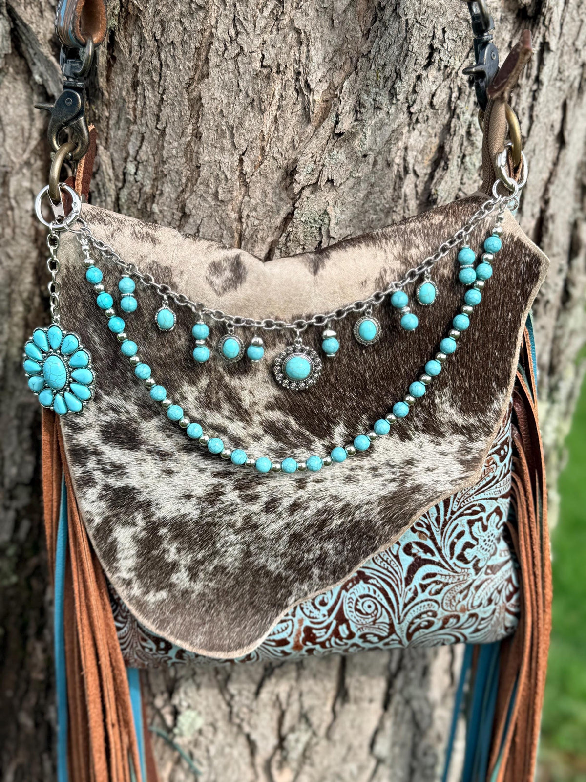 Alternate front view of brown and black cowhide crossbody bag with tan leather fringe against tree trunk with three-strand Western boho turquoise and silver purse chain showing silver cable chain with nine dangling turquoise cabochon cluster charms and crystal rhinestone concho pendant, turquoise round bead strand with silver rondelle spacers, and large turquoise flower concho on left side hardware, Western boho handbag accessory