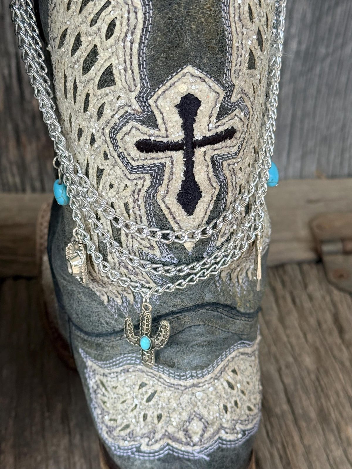 Close-up of dark gray distressed cross-embroidered cowboy boot shaft with three-strand silver chain lei boot bracelet featuring turquoise oval bead accents and silver cactus charm with turquoise stone centerpiece