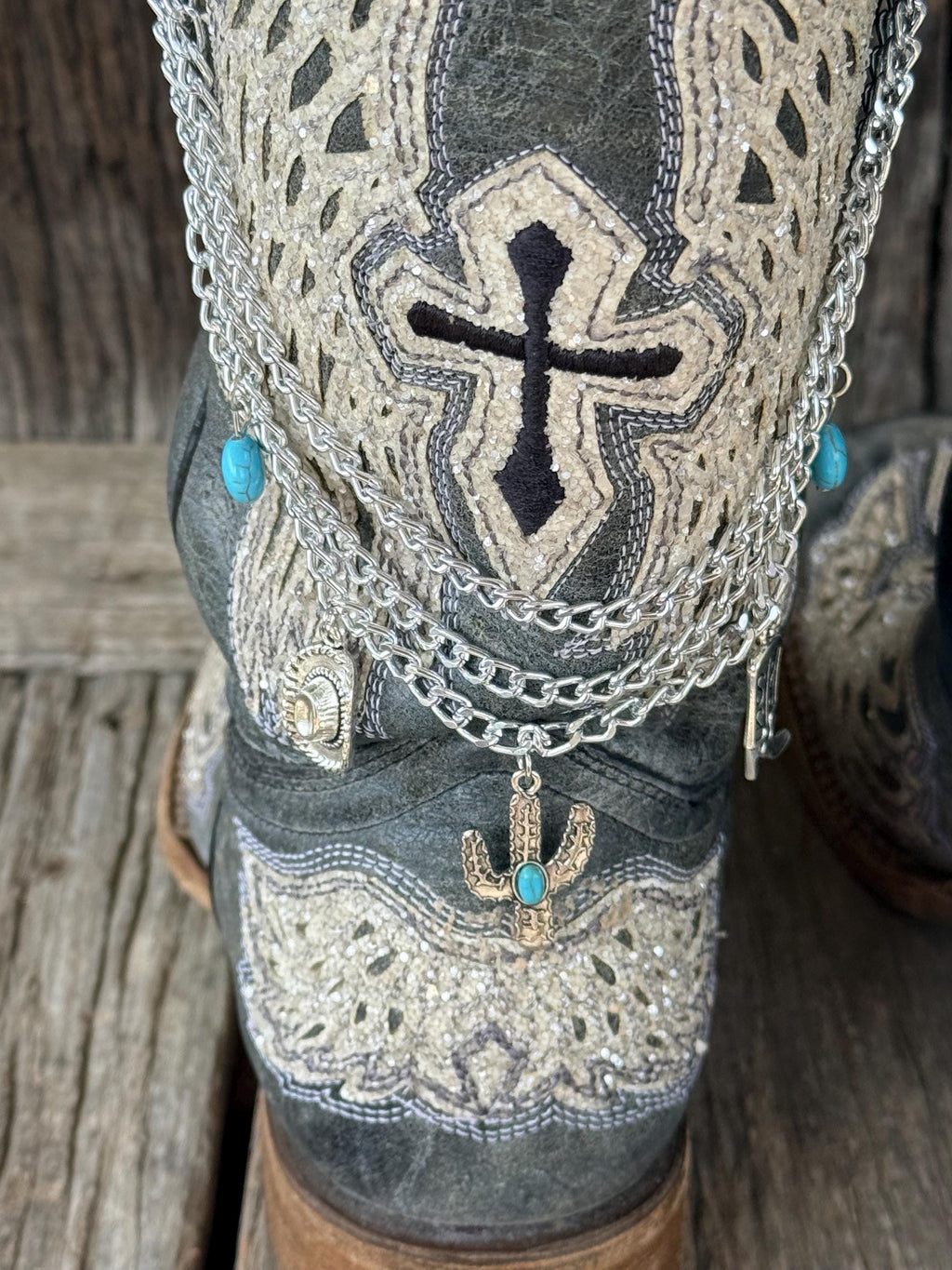 Close-up of dark gray distressed cross-embroidered cowboy boot with three-strand silver chain lei boot bracelet showing turquoise oval bead accents, silver cactus charm with turquoise stone, and silver cowboy hat charm