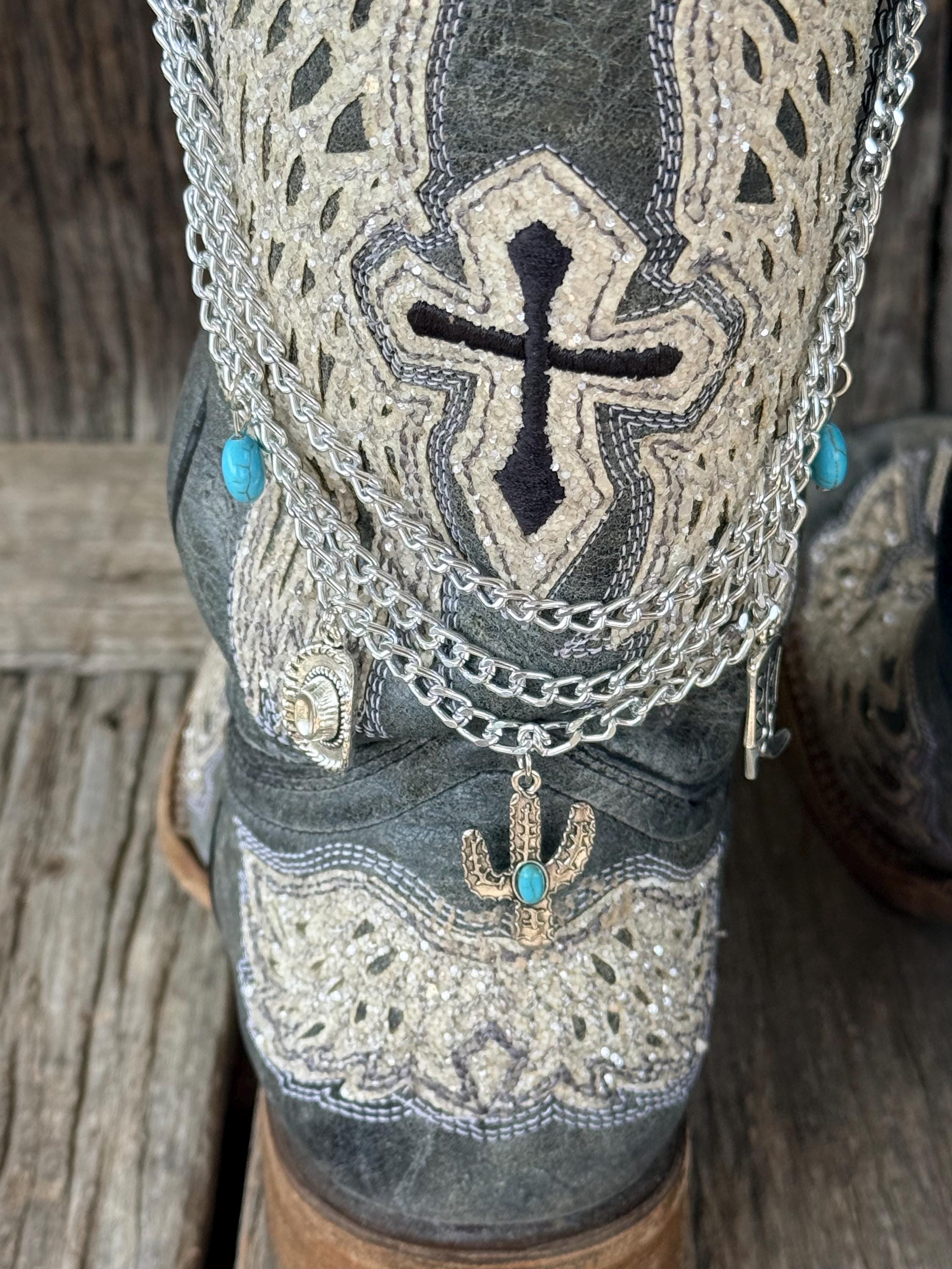 Close-up of dark gray distressed cross-embroidered cowboy boot with three-strand silver chain lei boot bracelet showing turquoise oval bead accents, silver cactus charm with turquoise stone, and silver cowboy hat charm