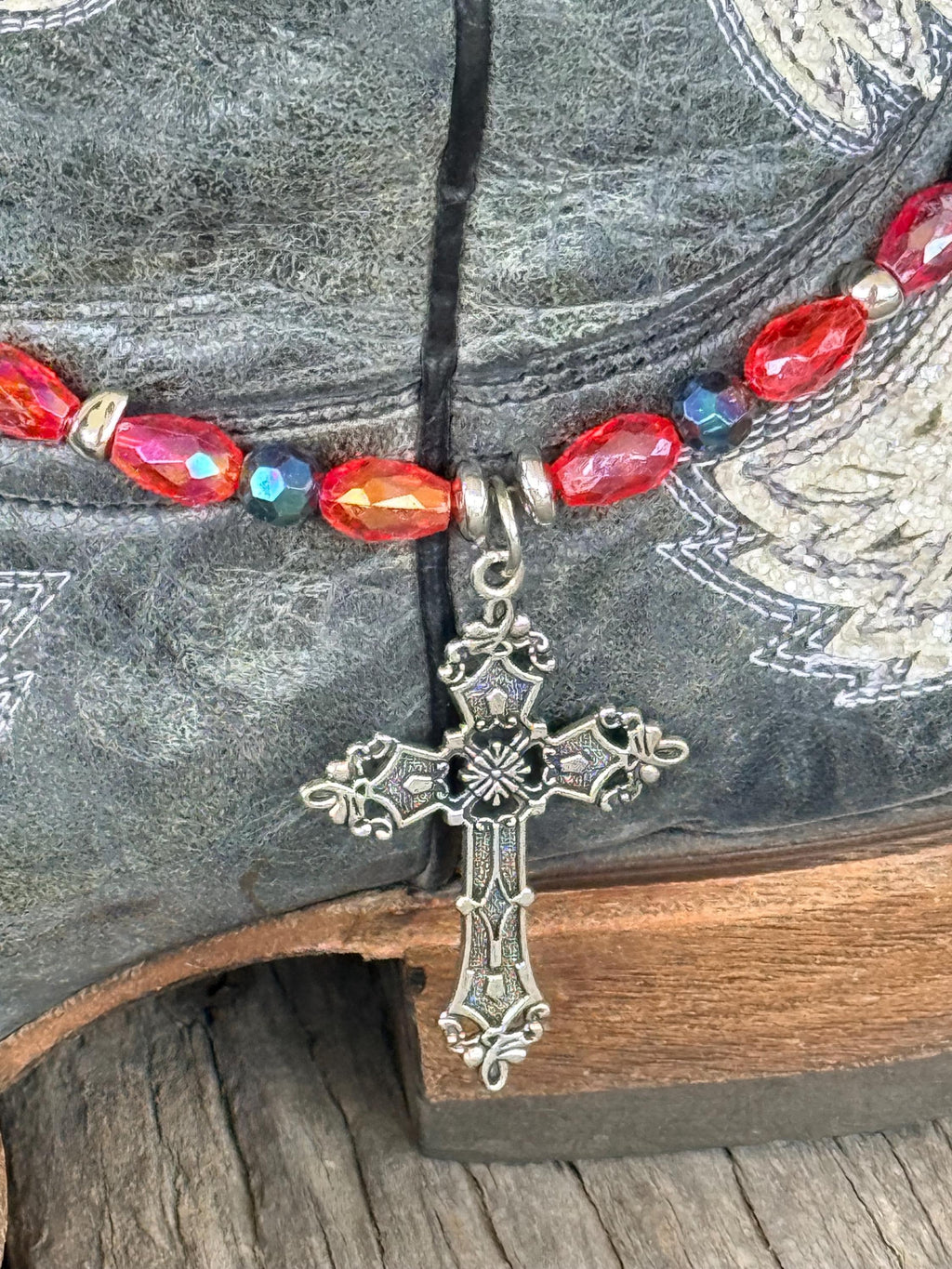 Close-up of dark gray distressed angel wing cowboy boot shaft with red AB oval crystal and dark teal iridescent bead boot bracelet and large ornate silver filigree cross pendant, faith-forward Western boot bling