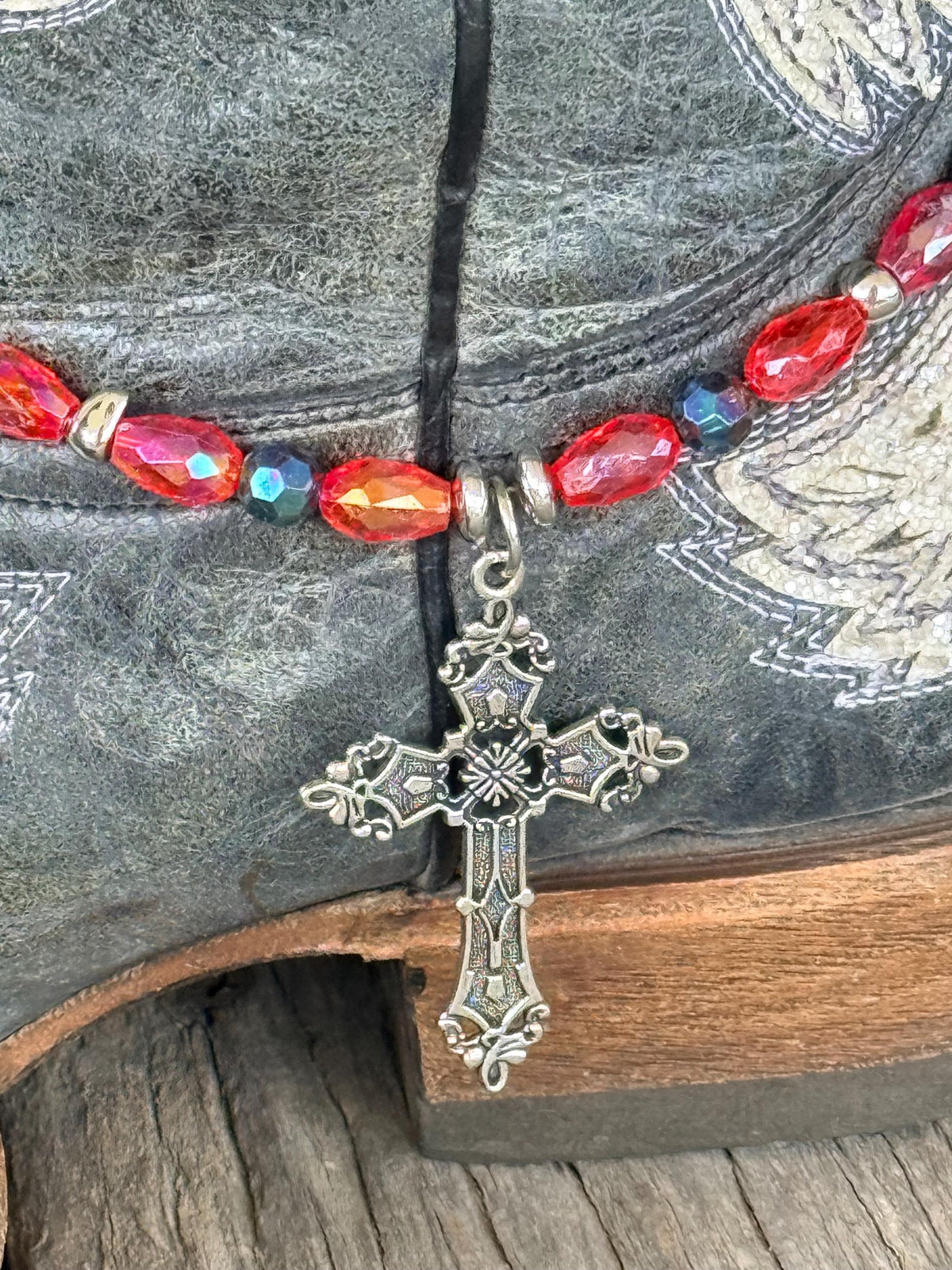 Close-up of dark gray distressed angel wing cowboy boot shaft with red AB oval crystal and dark teal iridescent bead boot bracelet and large ornate silver filigree cross pendant, faith-forward Western boot bling