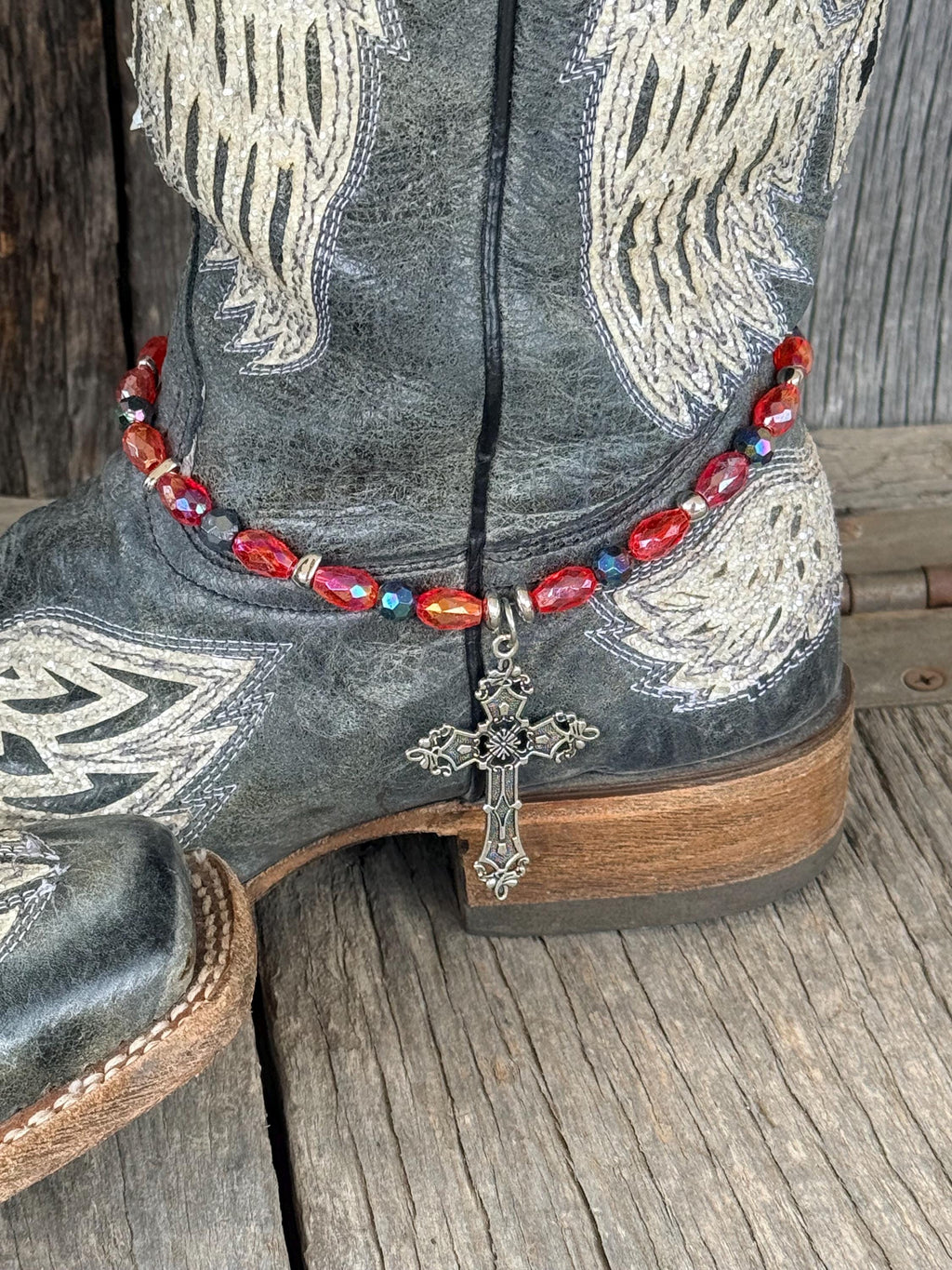 Front view of dark gray distressed angel wing cowboy boot with red AB oval crystal and dark teal iridescent bead boot bracelet featuring large ornate silver filigree cross pendant, Western boot bling