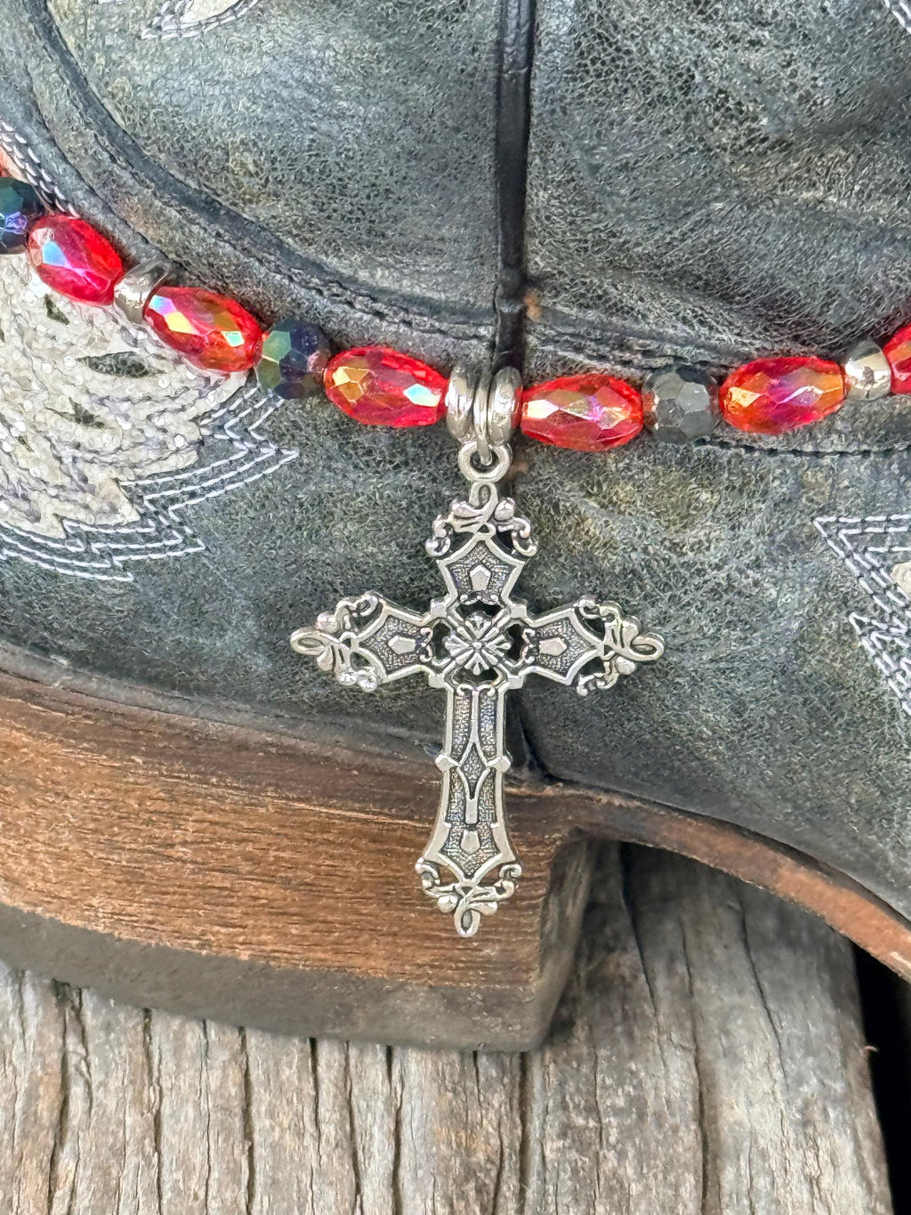 Macro close-up of large ornate silver filigree cross pendant with scrollwork and fleur-de-lis tips on red AB oval crystal and dark teal iridescent bead boot bracelet, on dark gray distressed angel wing cowboy boot