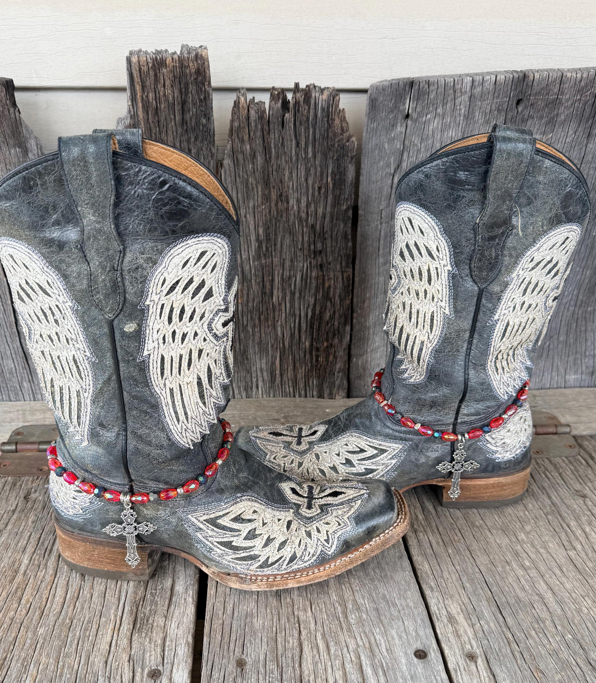 Front view of pair of dark gray distressed angel wing embroidered cowboy boots on weathered wood planks wearing red AB oval crystal and dark teal iridescent bead boot bracelets with large ornate silver filigree cross pendant, Western boot bling set of 2