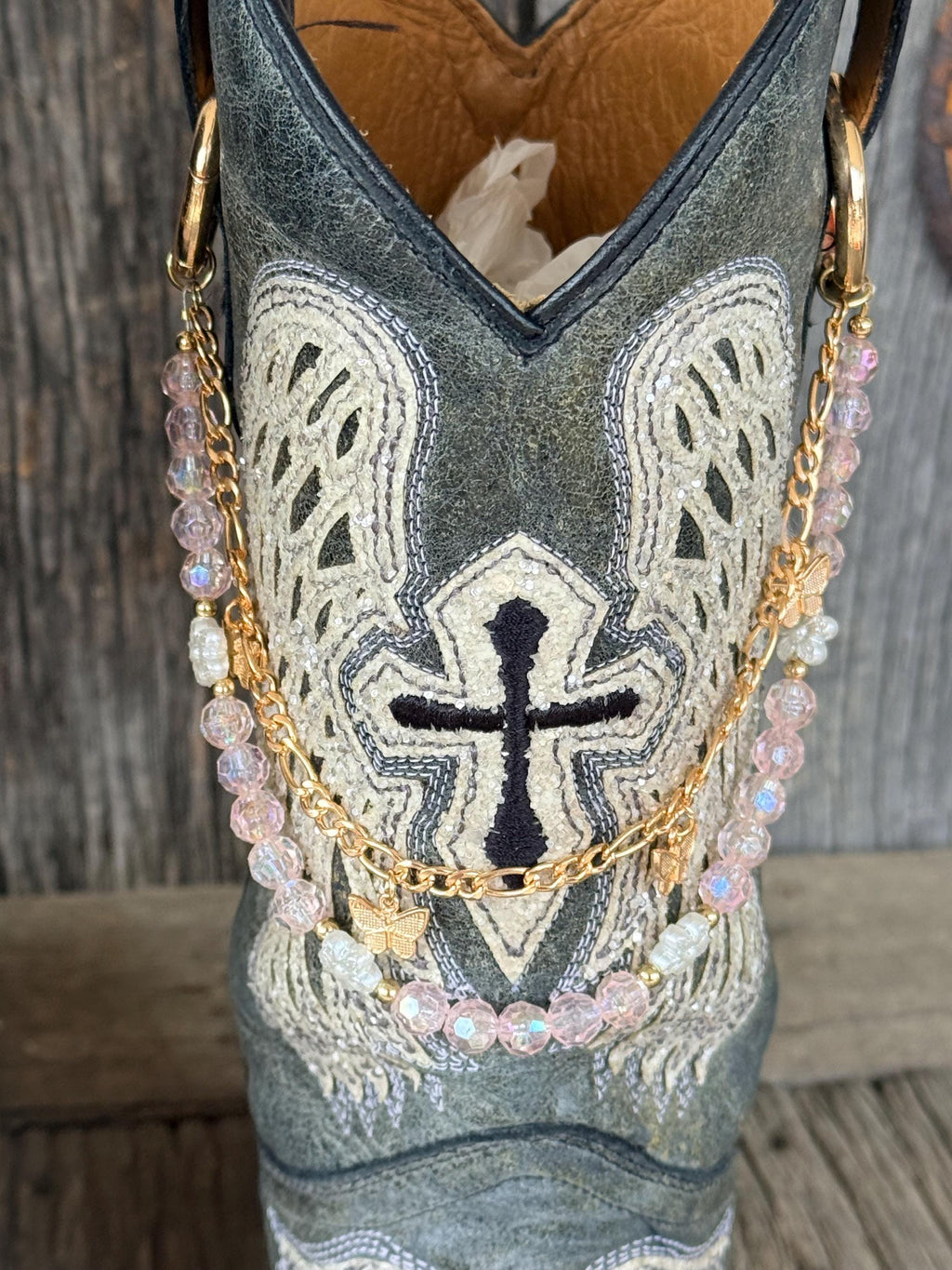 Front view of grey cross embroidered cowboy boot with layered gold link chain and iridescent pink crystal bead boot bracelet featuring gold butterfly charm pendant