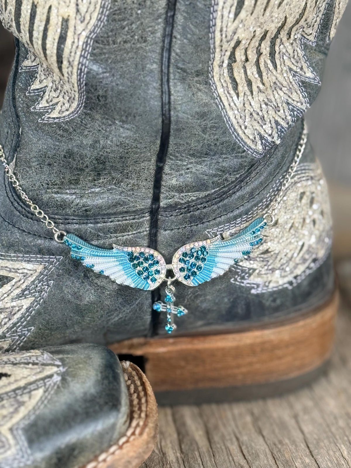 A close-up of a western-style boot with distressed gray leather and silver embroidery, featuring a winged pendant boot bracelet with turquoise blue rhinestones and a dangling cross charm.