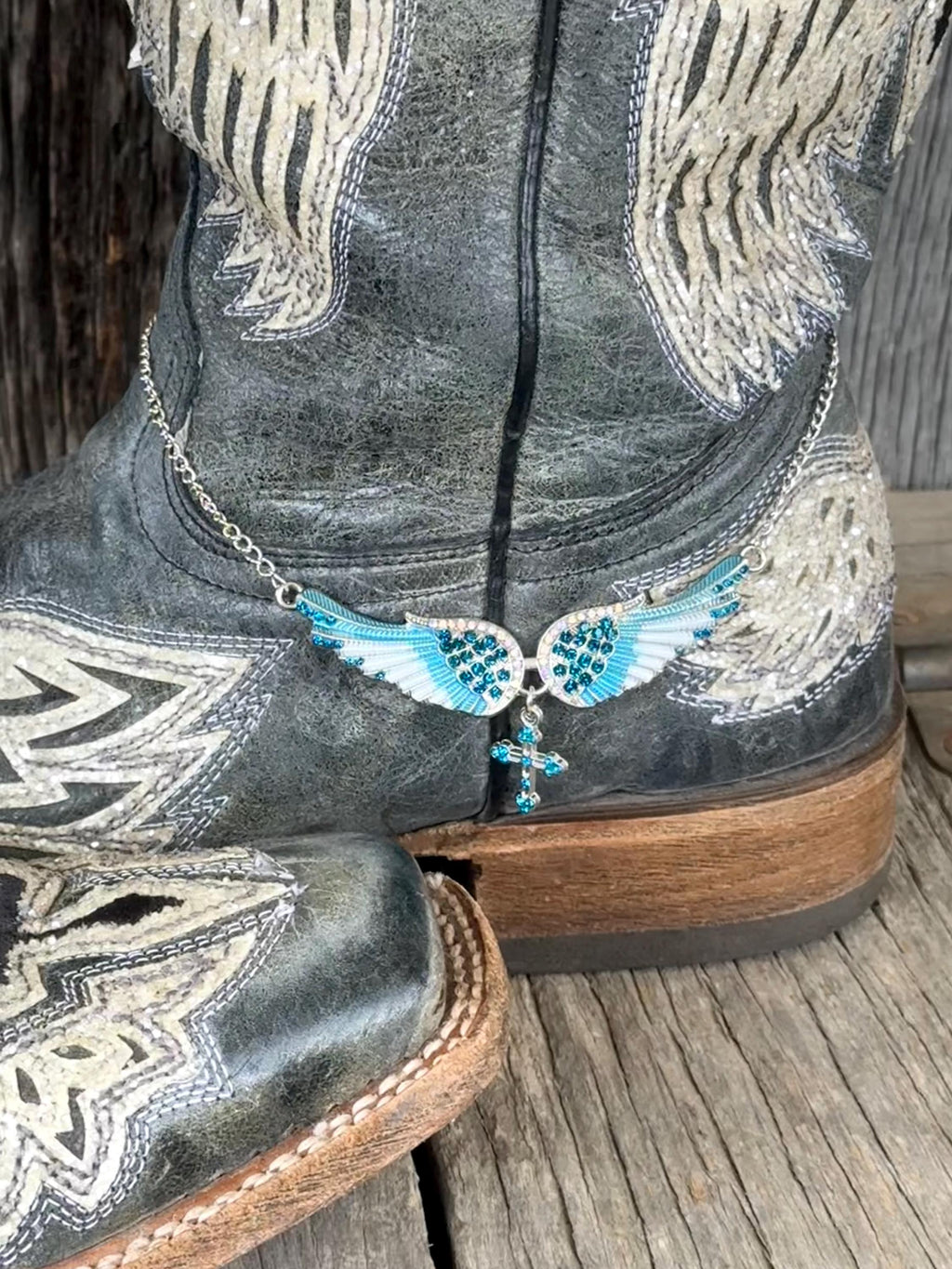 A close-up of a western boot with distressed charcoal gray leather, white embroidery, and a silver winged cross charm with turquoise blue accents resting near the ankle.