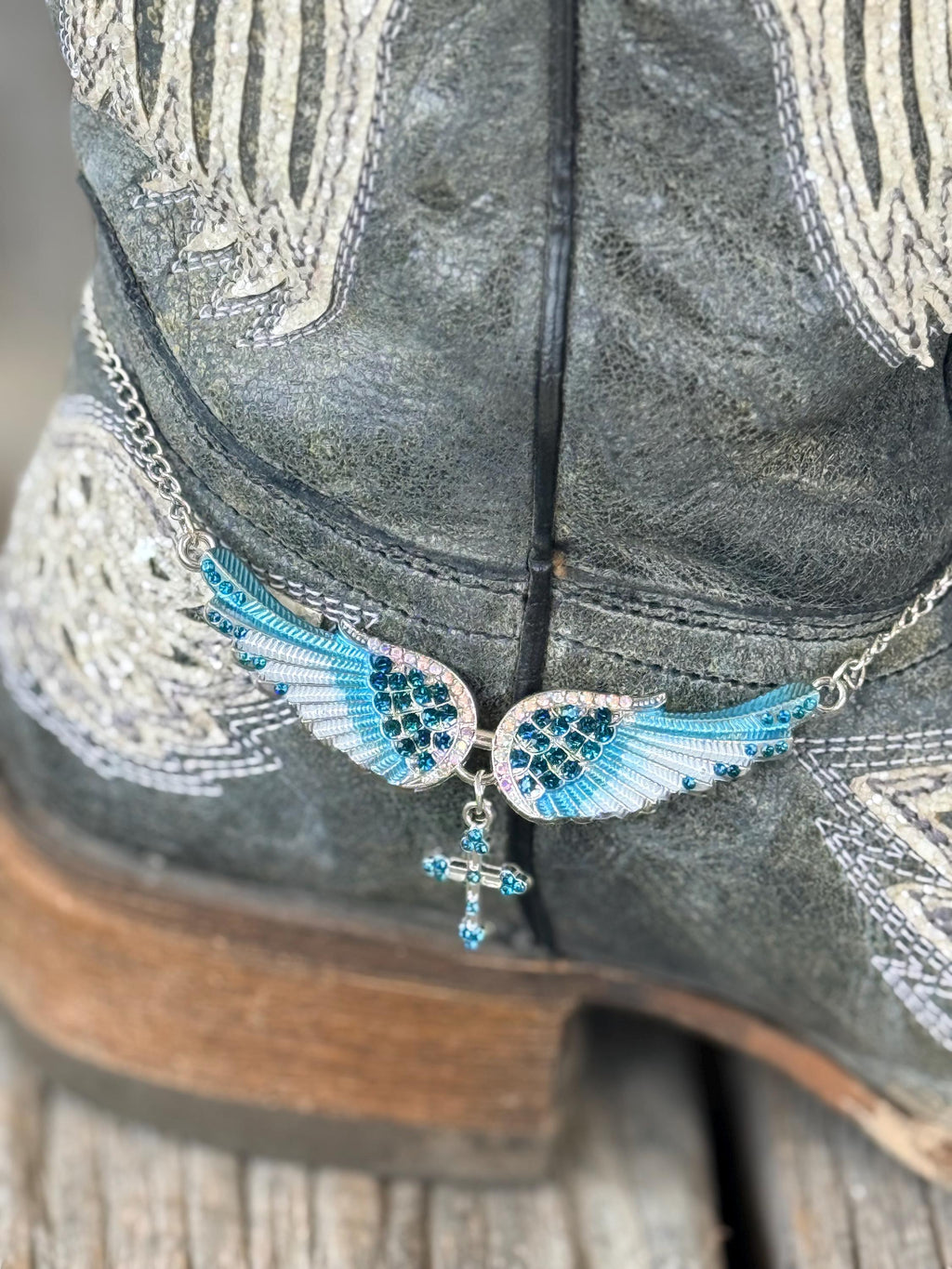 A close-up of a western boot in distressed charcoal gray with silver embroidery, featuring a decorative ankle charm with blue and turquoise wing motifs and a dangling cross on a chain.