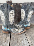 A pair of distressed gray leather cowboy boots with large white wing embroidery and turquoise wings & cross embellishments, displayed on a rustic wooden plank surface.