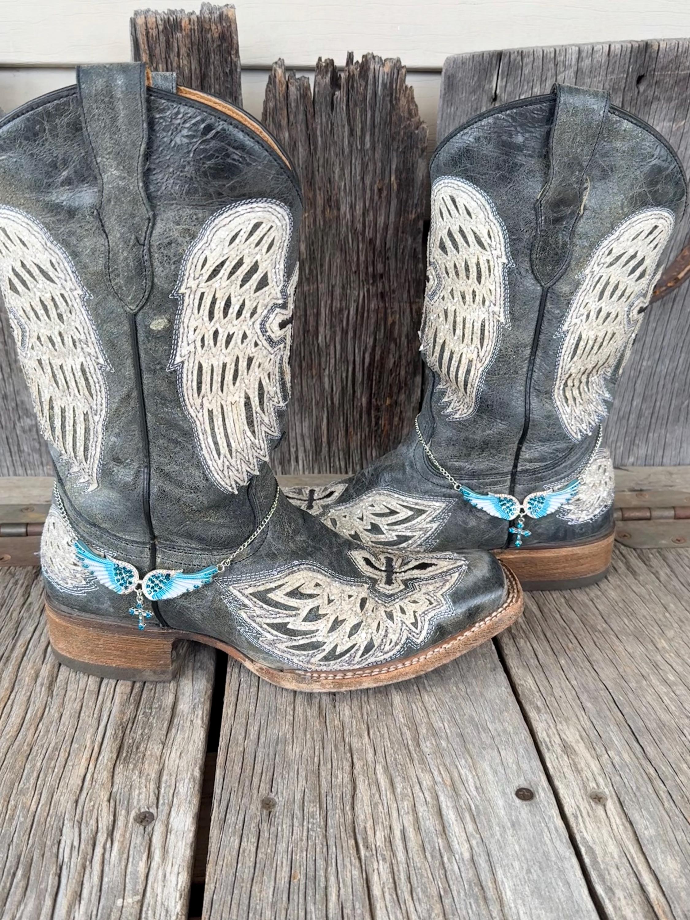 A pair of distressed gray leather cowboy boots with large white wing embroidery and turquoise wings & cross embellishments, displayed on a rustic wooden plank surface.
