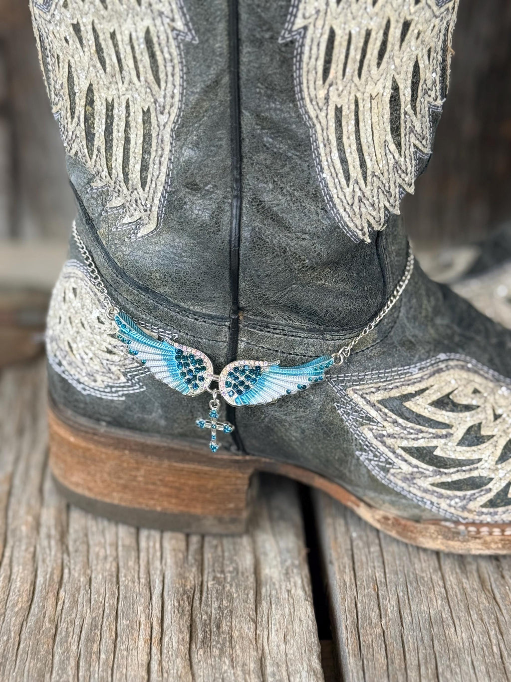 A close-up of a gray western boot with white embroidered wing patterns and a silver chain ankle bracelet featuring blue and white wings with a dangling cross charm, resting on a rustic wooden surface.
