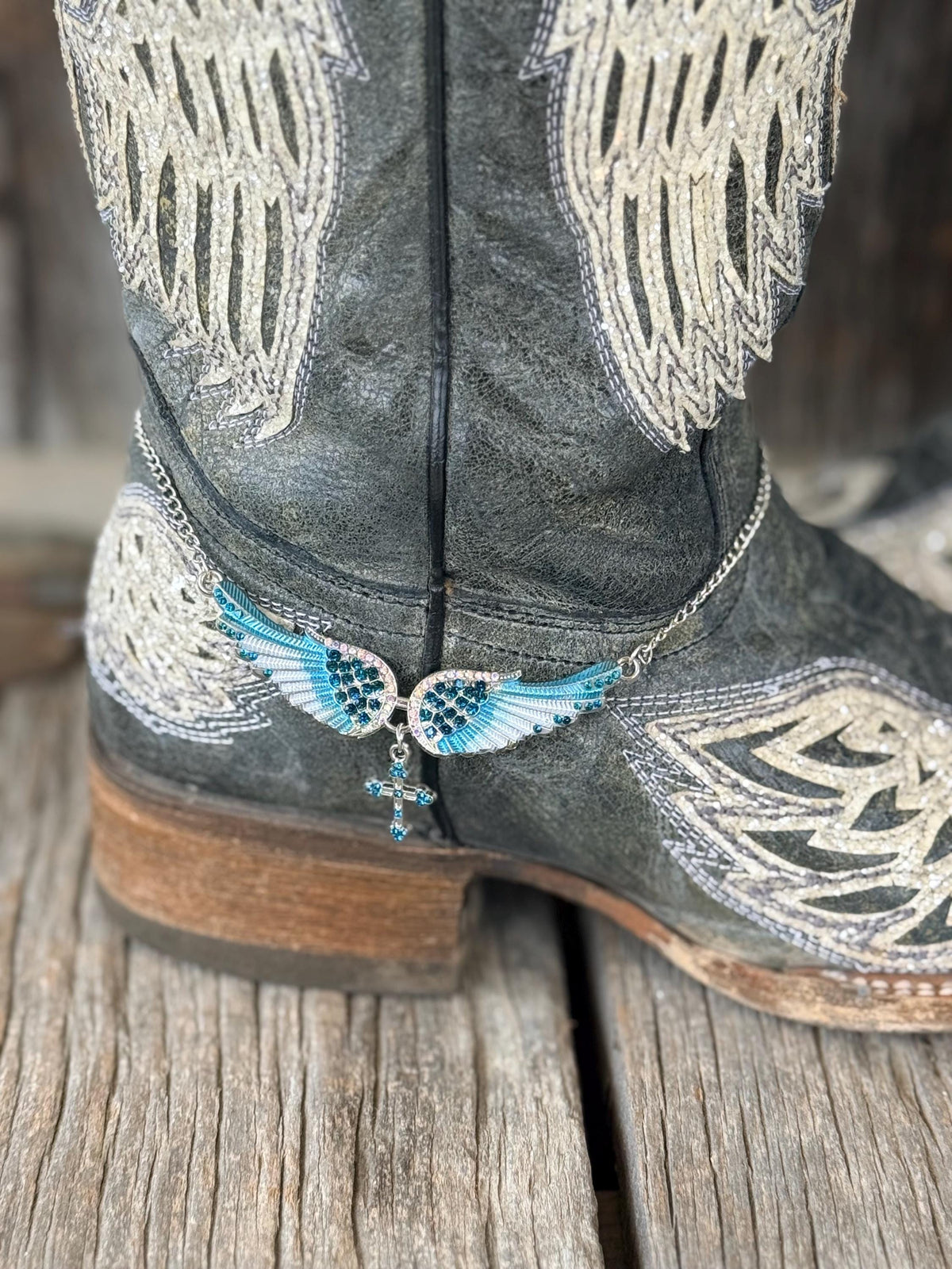 A close-up of a gray western boot with white embroidered wing patterns and a silver chain ankle bracelet featuring blue and white wings with a dangling cross charm, resting on a rustic wooden surface.