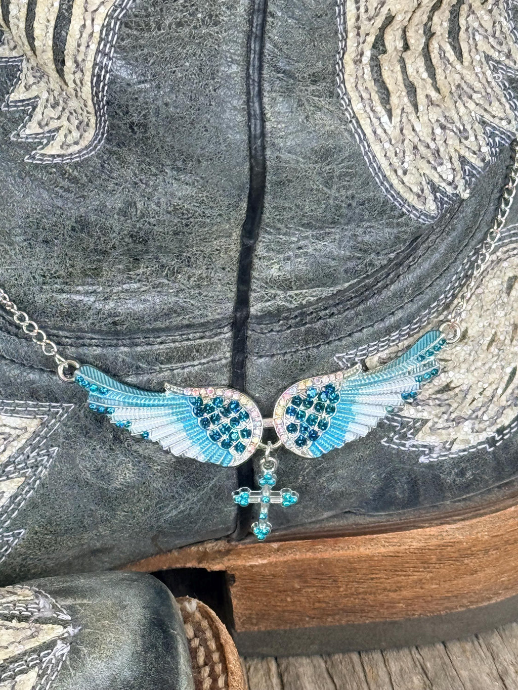 A close-up of a western-style necklace with a wing and cross pendant, featuring turquoise blue rhinestones on a silver chain, draped over a distressed gray leather cowboy boot.