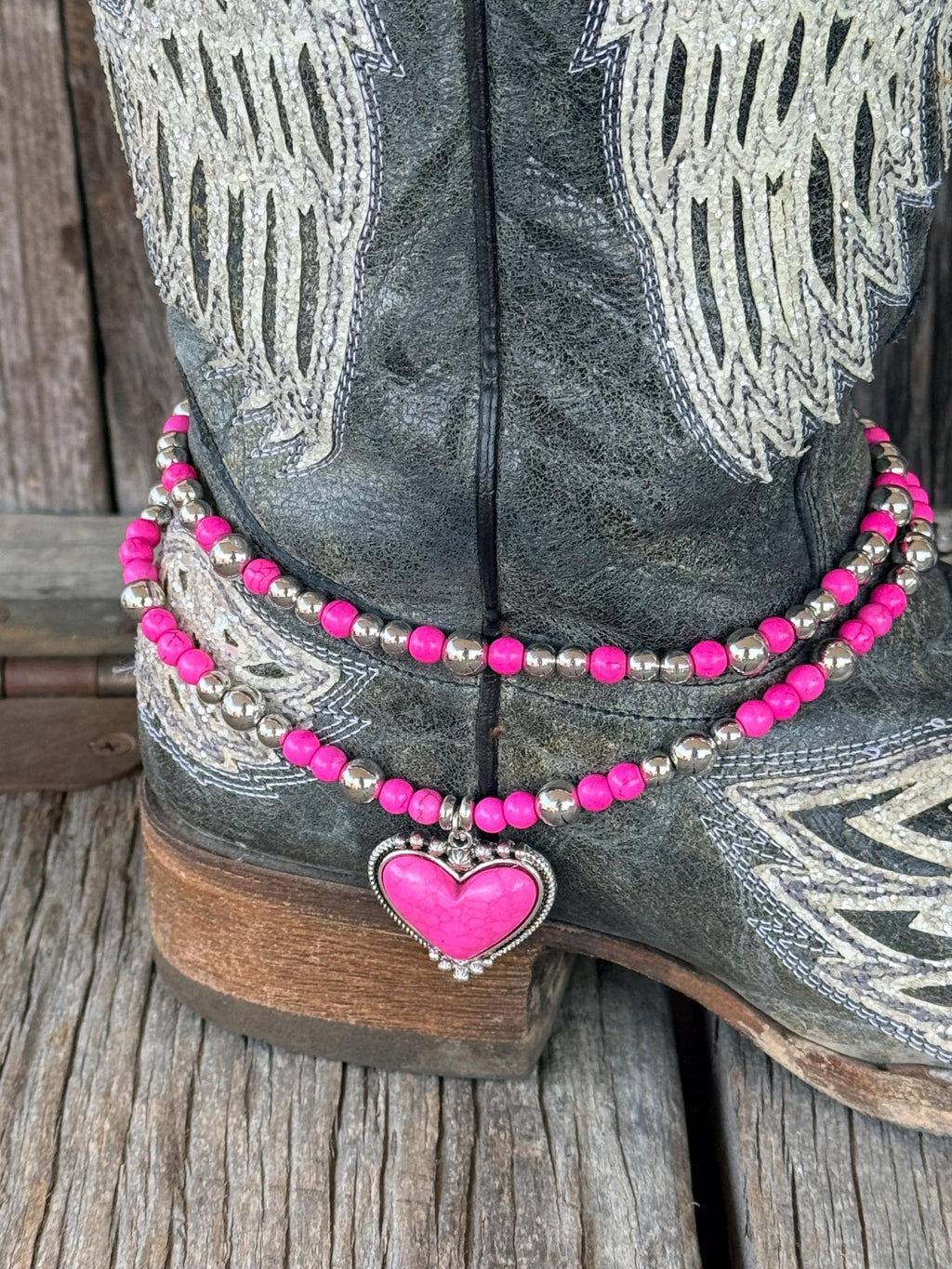 Close-up of black distressed angel wing cowboy boot ankle with two-strand hot pink round bead and silver ball spacer boot bracelet chain featuring large hot pink stone heart pendant set in decorative silver rope-edge bezel frame