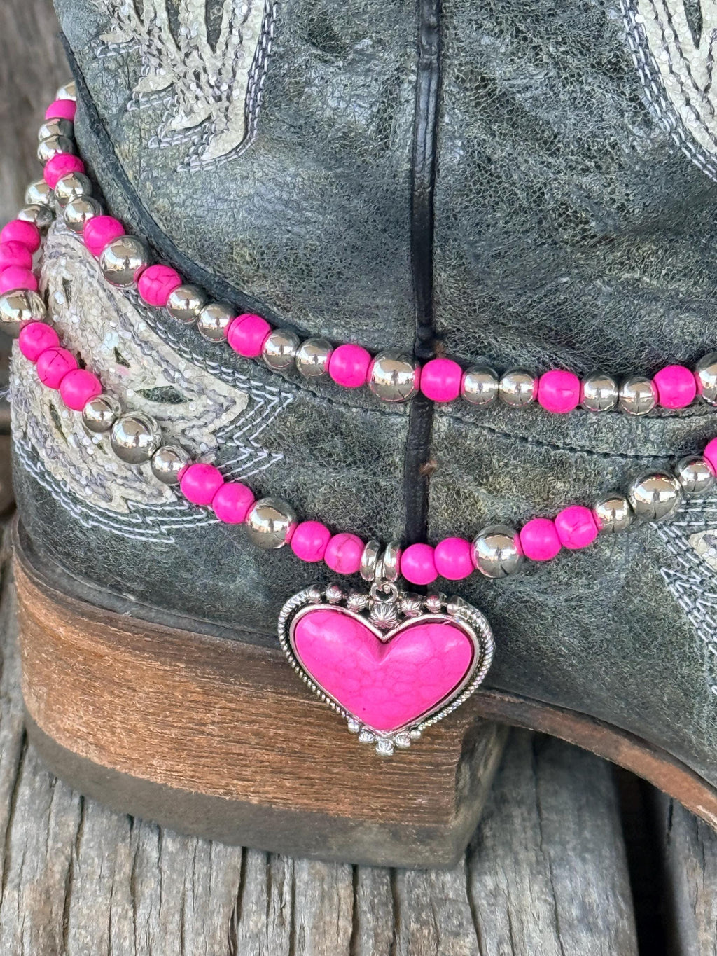 Close-up of black distressed cross and scroll embossed cowboy boot with two-strand hot pink round bead and silver ball spacer boot bracelet chain and large hot pink stone heart pendant in silver rope-edge bezel frame