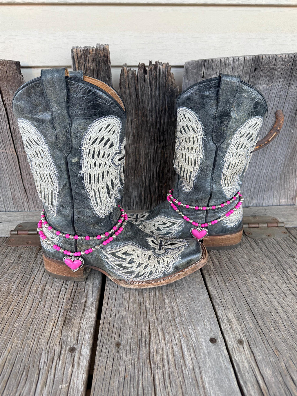 Front view of pair of black distressed angel wing and cross laser-cut cowboy boots on weathered wood planks wearing two-strand hot pink round bead and silver ball spacer boot bracelet chains with large hot pink stone heart pendant in decorative silver rope-edge bezel frame, Western boot bling set of 2