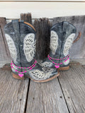 Front view of pair of black distressed angel wing and cross laser-cut cowboy boots on weathered wood planks wearing two-strand hot pink round bead and silver ball spacer boot bracelet chains with large hot pink stone heart pendant in decorative silver rope-edge bezel frame, Western boot bling set of 2