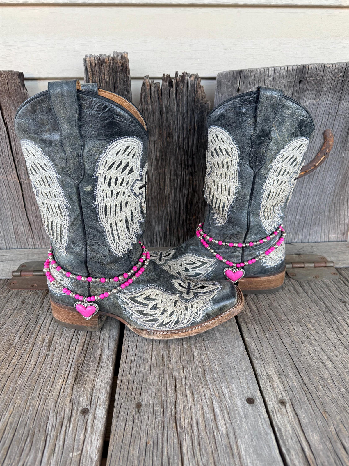Front view of pair of black distressed angel wing and cross laser-cut cowboy boots on weathered wood planks wearing two-strand hot pink round bead and silver ball spacer boot bracelet chains with large hot pink stone heart pendant in decorative silver rope-edge bezel frame, Western boot bling set of 2
